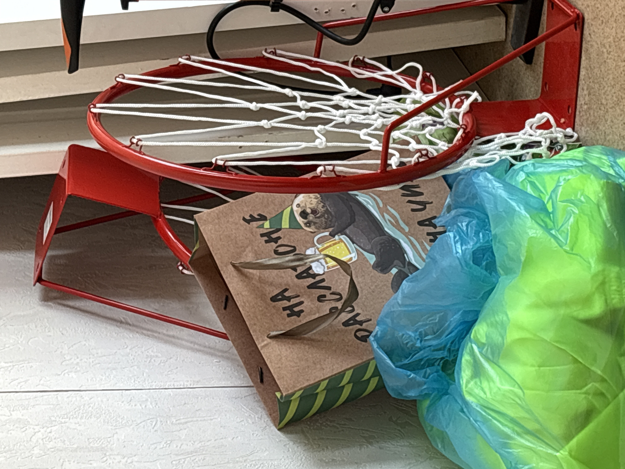 A close-up shot shows a red basketball hoop with a white net placed on top of a brown gift bag featuring an illustration of an otter wearing a party hat and holding a beer, along with Russian text. The gift bag has green and yellow stripes at the bottom. To the right of the gift bag is a crumpled blue and green plastic bag. The scene appears to be indoors on a white tiled floor. There is no information to determine that this is in Izhevsk, Russia.