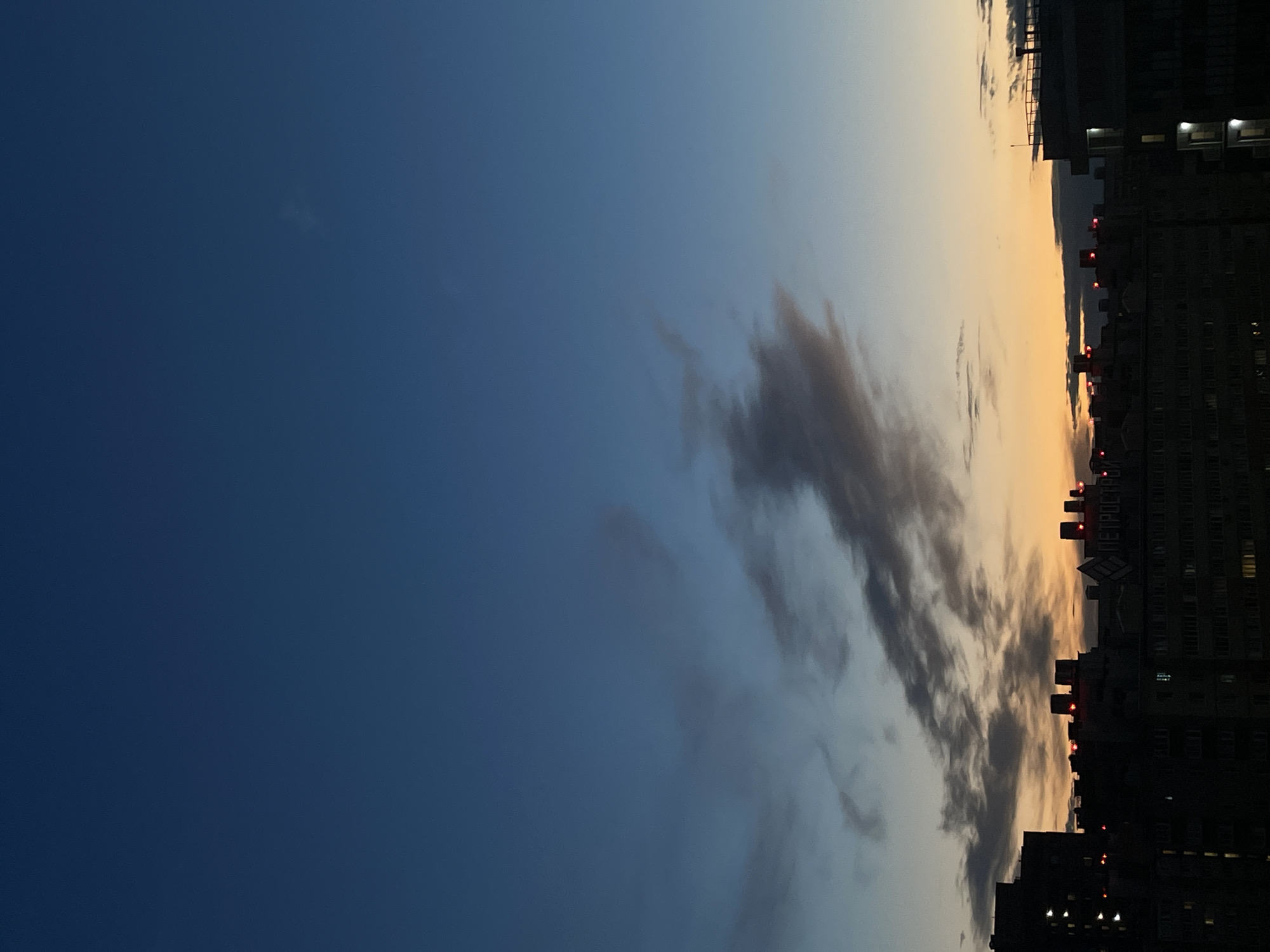 This is a vertically oriented image of a city skyline at dusk. The sky transitions from a deep blue on the left to a pale orange and yellow on the right, indicating the setting sun. Wispy clouds are visible, some dark and silhouetted against the lighter sky.
The foreground is dominated by the dark silhouettes of several buildings. Windows are visible in some of the buildings, with a few lit from within, suggesting people are inside. Red lights are visible atop some of the structures, possibly indicating hazards or air traffic control. A sign with Cyrillic text is partially visible on one of the buildings.
The image captures a quiet, urban atmosphere as day turns into night. The mood is serene and contemplative, with the warm glow of the sunset juxtaposed against the encroaching darkness of the city. The time of day is clearly dusk, and the weather appears clear apart from the scattered clouds. The location is indicated as Russia due to the Cyrillic text. No people are directly visible in the frame.