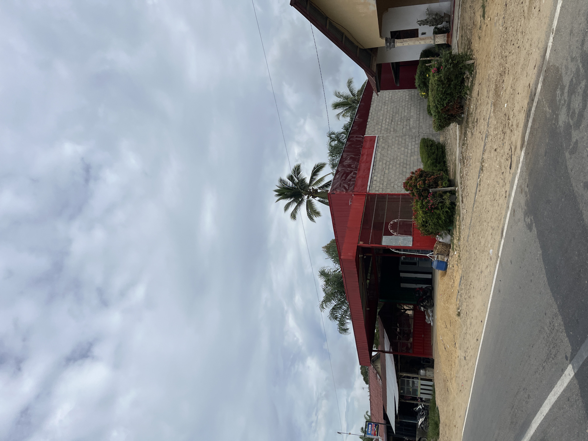 This image depicts a street-level view of a building with a red roof and white brick exterior, situated in Sukaramai Dua, Indonesia. The building appears to be a commercial establishment, with open storefronts and various objects visible inside, including what looks like motorcycles parked under a covered area. A paved road runs alongside the building, with a sandy, unpaved area between the road and the structure. Several well-maintained green bushes and some flowering plants are planted in front of the building. A prominent palm tree stands tall behind the building, reaching towards the overcast sky.
The weather is cloudy, suggesting it could be a warm, humid day, typical of tropical regions. There are no people clearly visible in the frame, and the scene appears to be at a standstill, with no obvious activity taking place. Power lines are visible stretching across the sky. On the far right of the image, a sign with blue and white text is partially visible, though the text is too blurry to be read clearly. The perspective of the photo is slightly tilted, giving it a dynamic feel. The overall impression is of a quiet, everyday scene in a rural or semi-urban Indonesian setting.