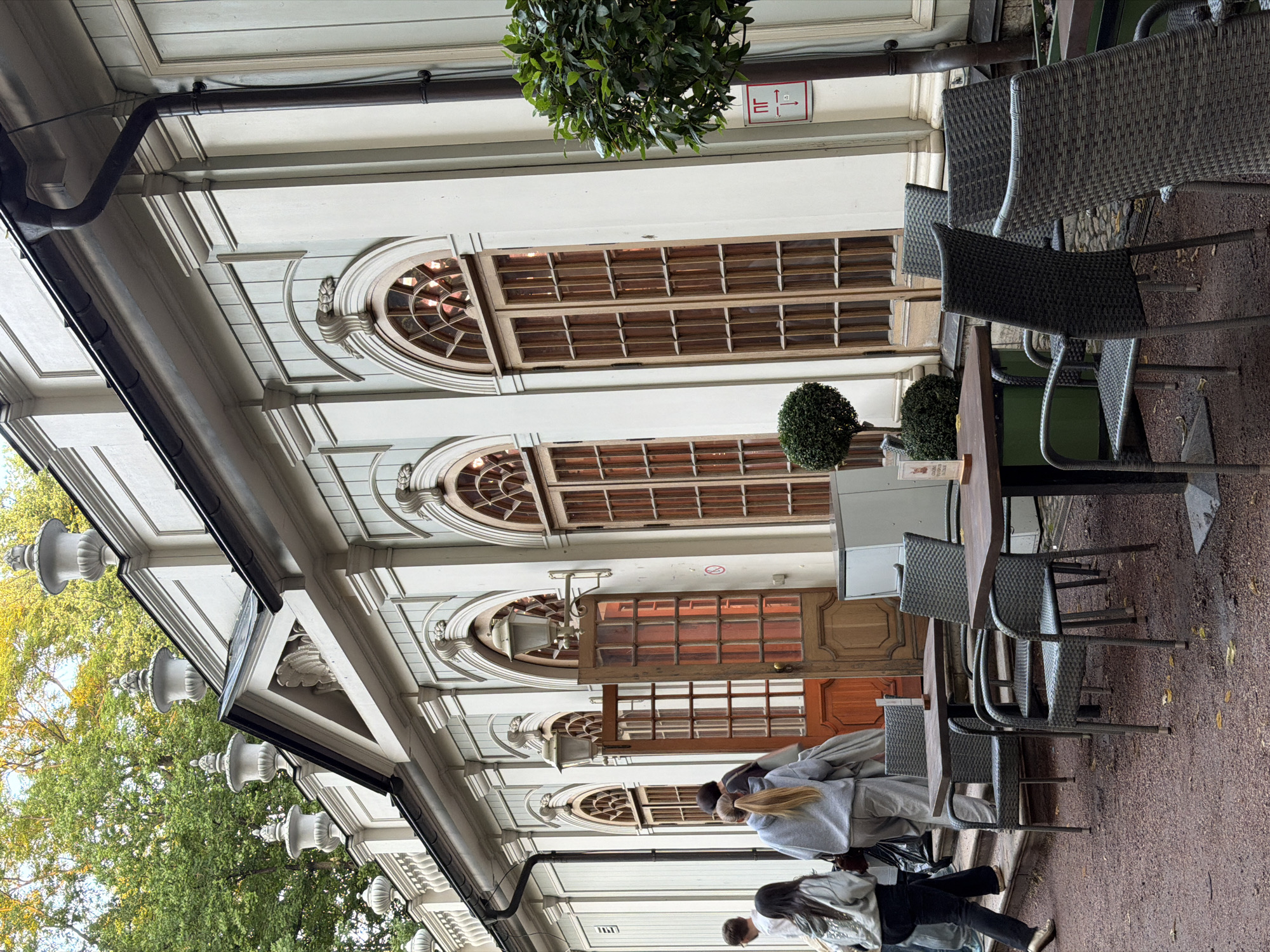 The image shows the exterior of a white building with classical architectural elements in Saint Petersburg, Russia. The building features arched windows with wooden panes, decorative moldings, and ornate urns along the roofline. A prominent dark brown gutter system is visible. In front of the building, there are several outdoor tables and chairs, suggesting a cafe or restaurant seating area. Two people, a woman in a grey hoodie and black pants, and another person in a grey hoodie and light-colored pants, are walking away from the building, with a third person partially visible behind them. The ground is covered in reddish gravel. To the right of the building, a small, well-maintained bush and a tree are present. A red and white sign with a pictogram is affixed to the wall near the tree. The scene appears to be a daytime shot, with dappled sunlight filtering through the trees in the background.