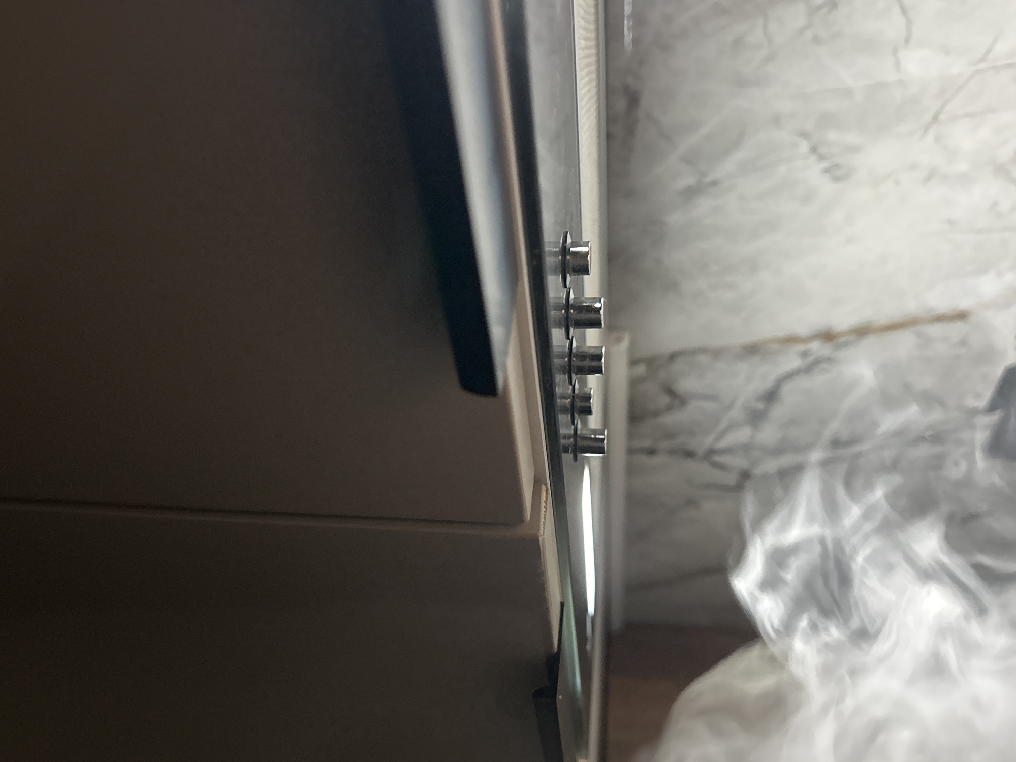 An upward-facing, eye-level, close-up shot shows the underside of a dark brown kitchen cabinet. Below the cabinet, a stainless steel range hood is visible, reflecting a bright light source. The range hood has several small, cylindrical metal components protruding downwards. The background consists of a marble-patterned backsplash with swirling grey and white veins. The overall scene suggests an indoor domestic environment, likely a kitchen.