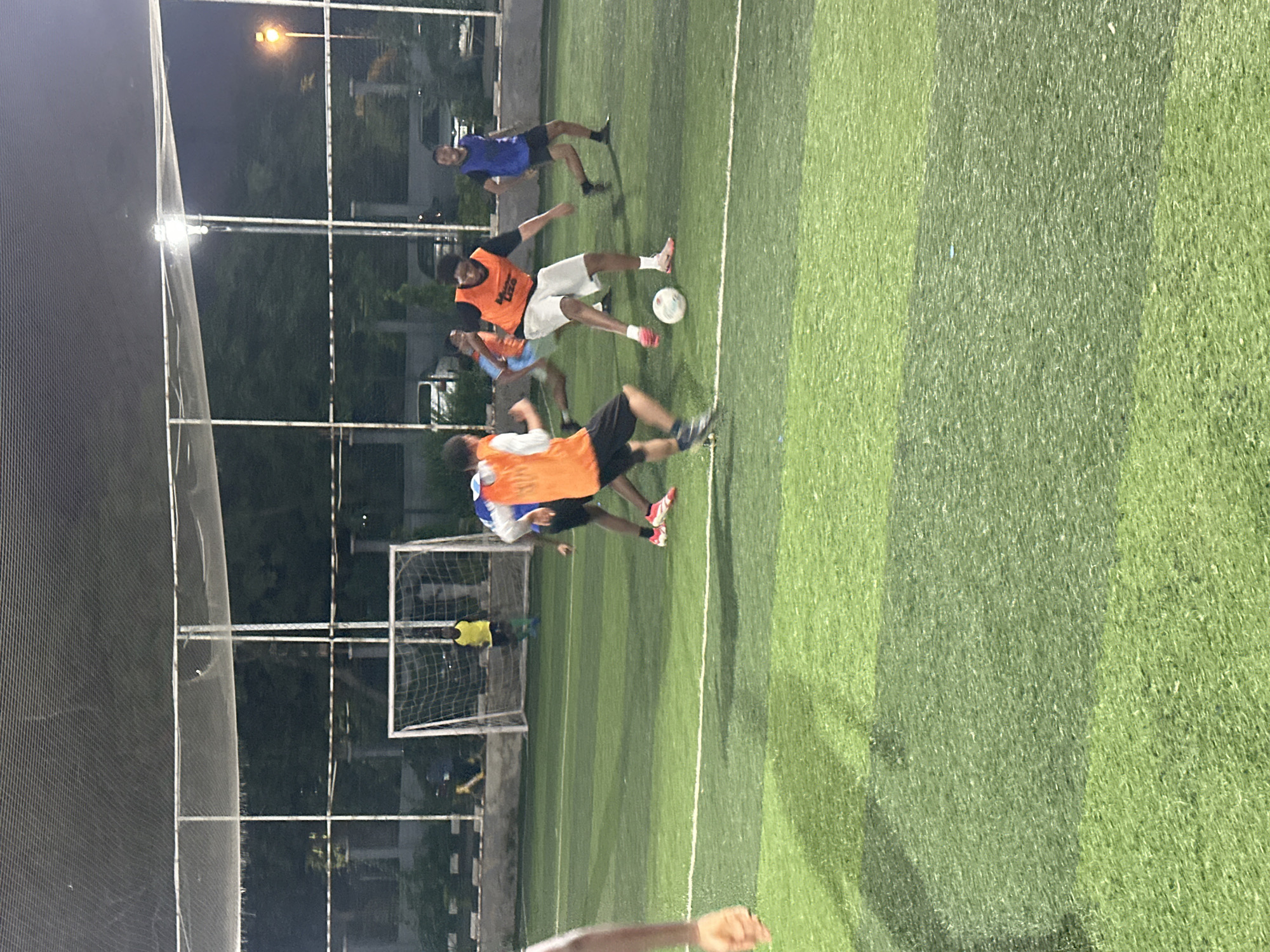 The image depicts a group of young men playing soccer on an artificial turf field at night. The field appears to be illuminated by artificial lighting, with floodlights visible in the background. The players are wearing various colored jerseys, some with orange bibs. One player in an orange bib is dribbling a soccer ball, while others are positioned around him, either defending or preparing to receive a pass. A soccer goal with a white net is visible in the background. The surrounding environment includes a fence, trees, and some parked vehicles, suggesting an urban setting. The overall impression is of an amateur soccer match taking place in a recreational area. The mention of Abuja, Nigeria, provides the geographical context for this scene.