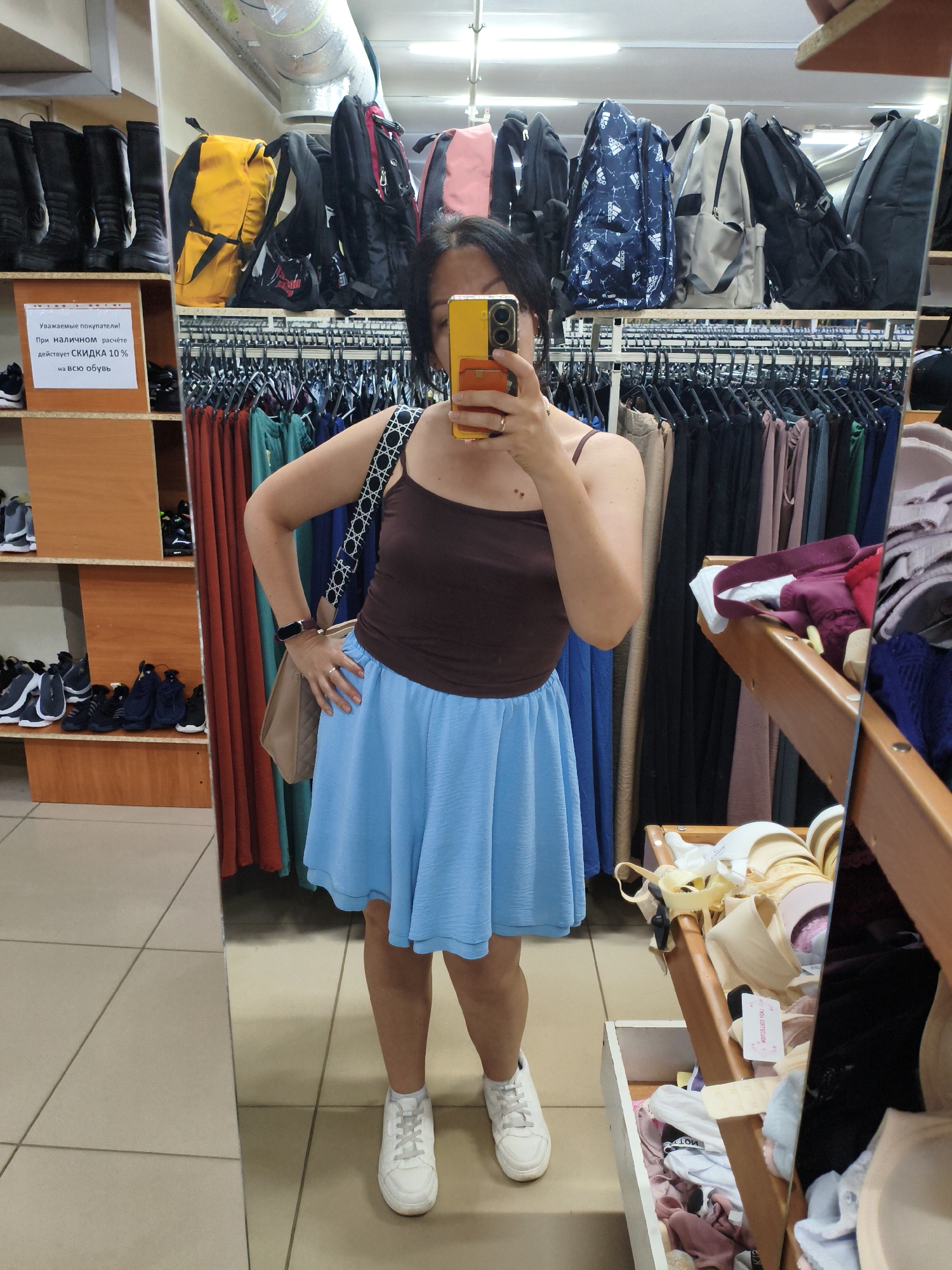 The image shows a woman taking a mirror selfie in a store. She is wearing a brown tank top, a light blue pleated skirt, and white sneakers. She is holding a yellow smartphone in her right hand, capturing her reflection. She has a light brown crossbody bag slung over her left shoulder and is wearing a smartwatch on her left wrist.
The store appears to be a clothing or shoe store. To the left of the woman, there are shelves displaying black boots and several backpacks on a higher shelf. Below the boots, more shelves hold various pairs of sneakers. Behind the woman, clothing racks are visible, filled with garments in various colors, including teal, red, and black. In the reflection of the mirror to the right, a display shelf is filled with bras and other lingerie items.
A sign in Russian is visible on the shelf to the left, indicating a 10% discount on all shoes. The floor is tiled with large, light-colored square tiles. The overall lighting suggests an indoor retail environment. The specific location in Orenburg, Russia, cannot be determined from the image itself.