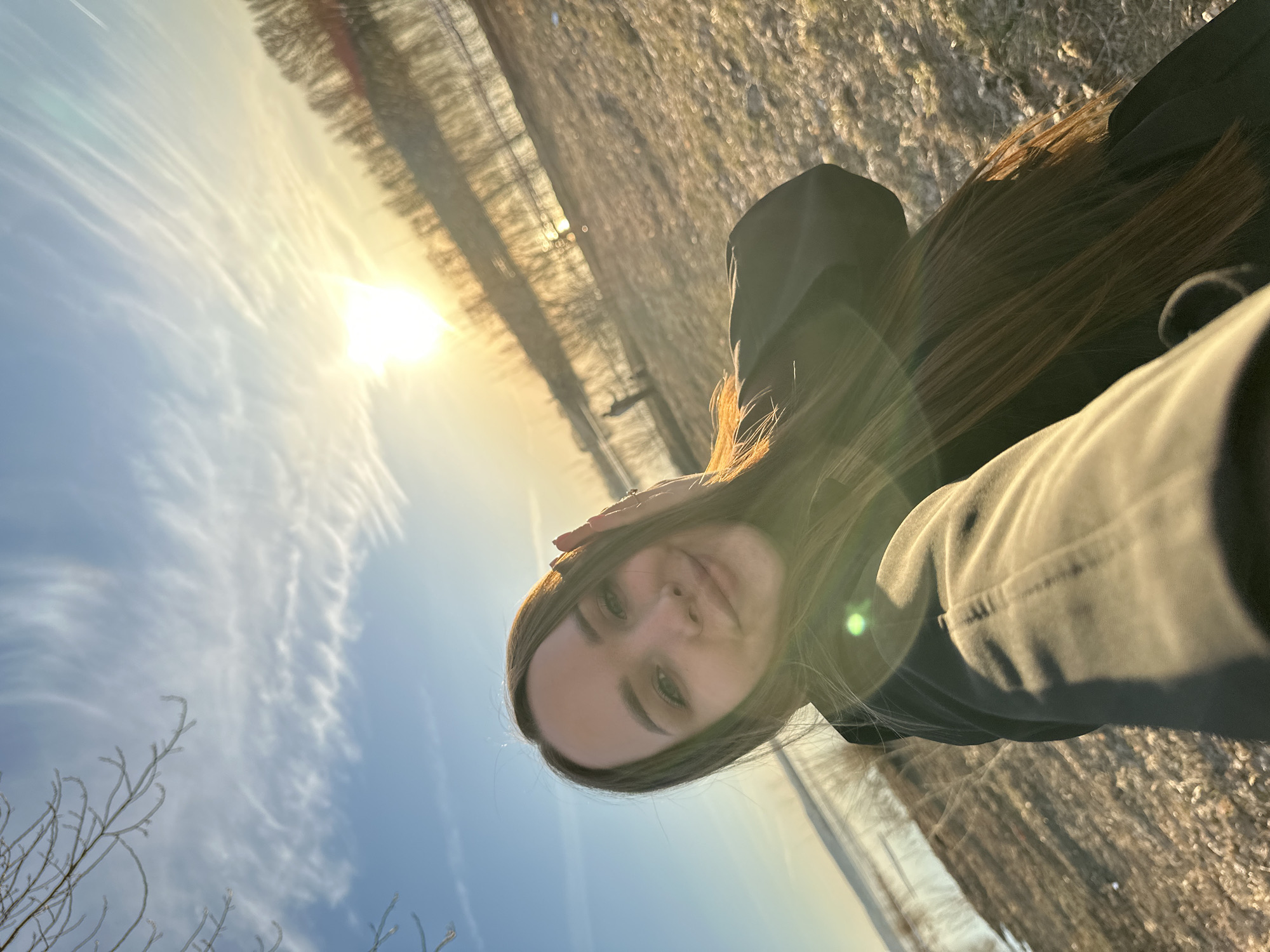 The image is a selfie featuring a young woman taken outdoors during what appears to be golden hour or sunset. The woman is positioned to the right side of the frame, looking directly at the camera with a neutral to slightly smiling expression. She has long, dark brown hair that is illuminated by the warm sunlight, and she is wearing dark clothing, possibly a hoodie or jacket. One hand is gently placed on the side of her head.
The background shows a natural outdoor landscape. A bright, low sun is a prominent feature on the left, casting a strong, warm light across the scene and creating noticeable lens flares. The sky is predominantly clear blue with some delicate, wispy white clouds. Below the horizon, a body of water, likely a river or lake, is visible, reflecting the light. The far bank of the water is lined with what appear to be bare trees or bushes, suggesting a cooler season such as late autumn, winter, or early spring. The ground in the immediate foreground is out of focus but appears to be dry earth or sparse vegetation. No visible text is present in the image.