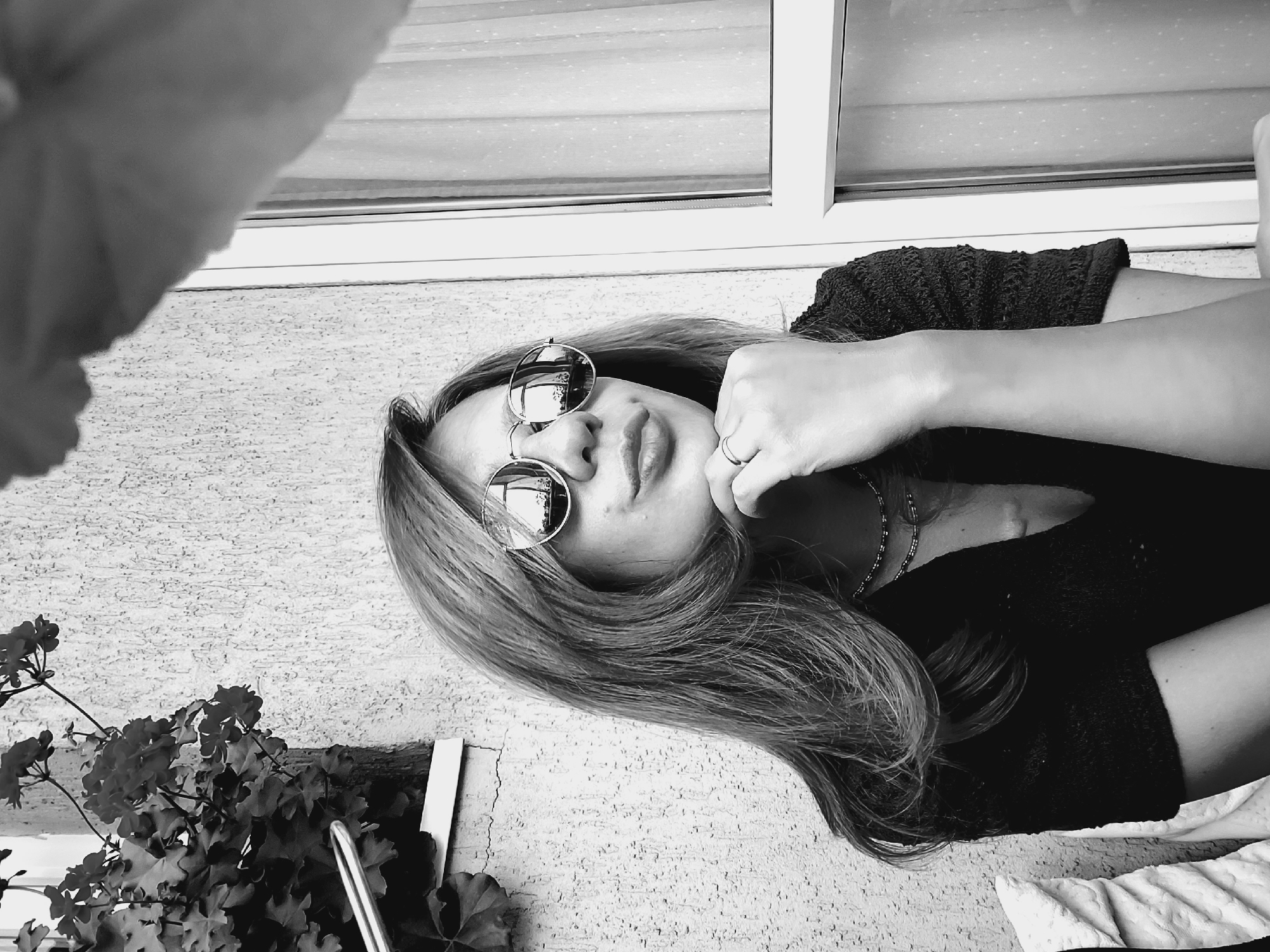 The black and white, low-angle, close-up, waist-up shot shows a woman lying down outside. She is wearing sunglasses and a dark, textured top. Her left hand is resting on her chin, and she has a ring on her right ring finger. A necklace is visible around her neck. Her long, blonde hair is spread out around her. Behind her is a textured wall and a window. To her left, some green plants with flowers are visible. The light suggests it is daytime. The image does not contain any visible text. The location context is Sofia, Bulgaria.