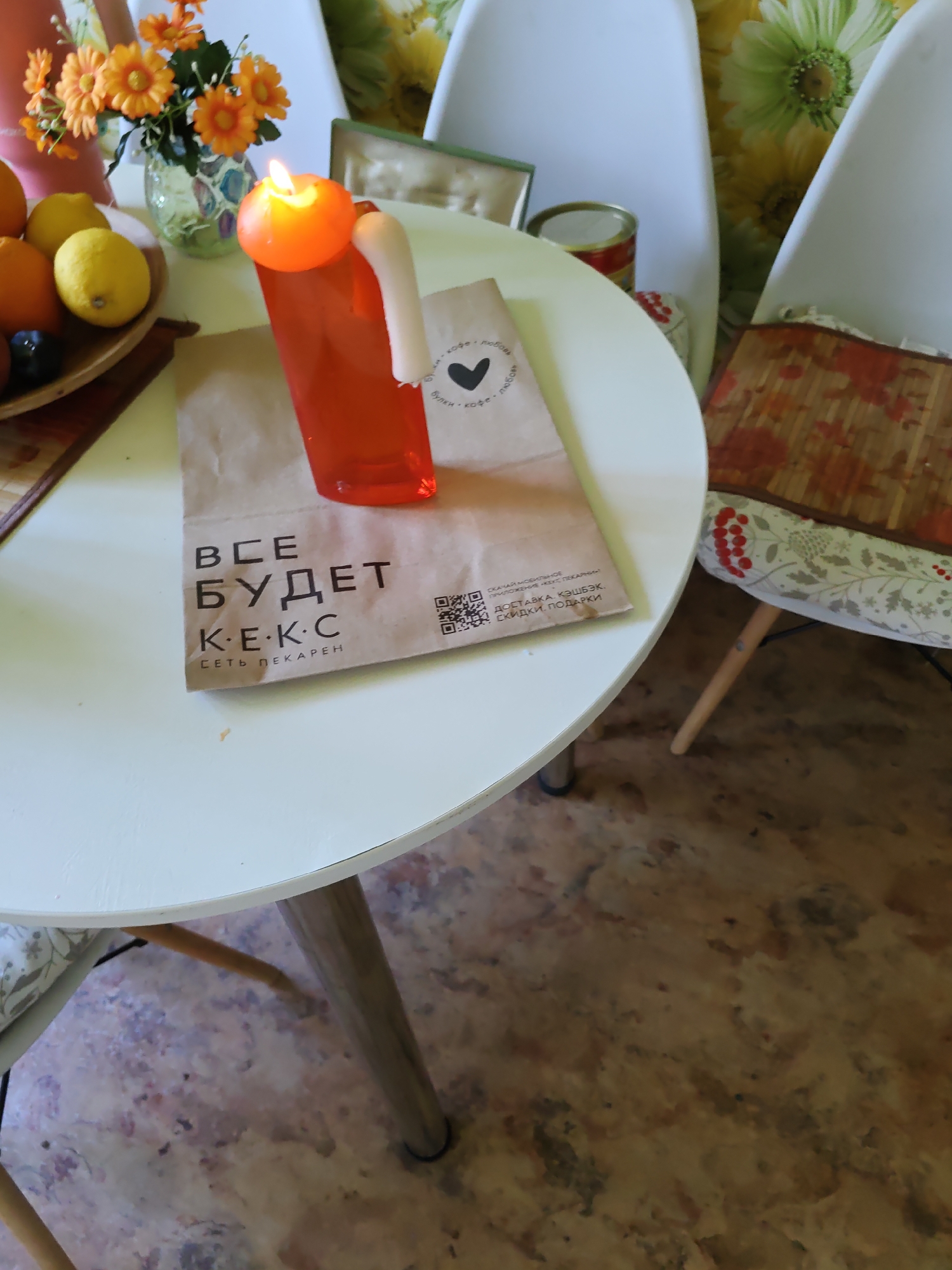 A table in a bright, indoor setting. On the table, a white paper bag with black text that reads "ВСЕ БУДЕТ КЕКС" and "СЕТЬ ПЕКАРЕН" is present. To the left of the bag, there is a small bowl containing fruit, including lemons and dark berries. Behind the fruit bowl, a vase with orange artificial flowers is visible. A lit orange candle in a white holder is placed on top of a transparent orange plastic container situated on the paper bag. In the background, two white chairs with patterned cushions are partially visible, and a wall with a floral wallpaper can be seen. The flooring appears to be a patterned linoleum. The scene is consistent with a domestic or casual cafe setting. The image does not provide any specific geographical indicators to determine a location in Rostov-on-Don, Russia.