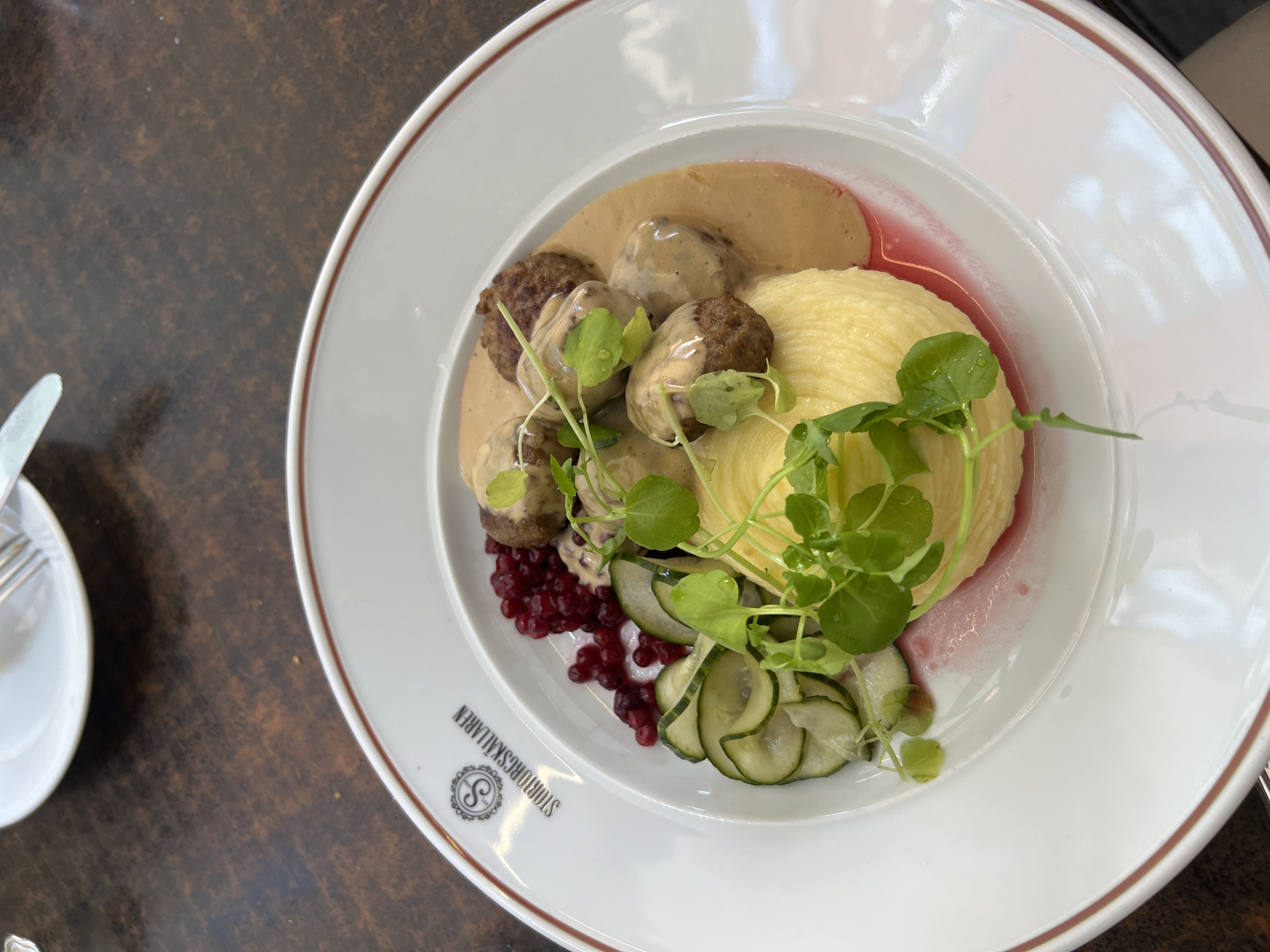 A plate of Swedish meatballs with mashed potatoes, lingonberries, and pickled cucumbers is presented on a wooden table. The plate has a red rim and the logo "STUREGÄLLAREN" is visible on the left side. The meatballs are coated in a creamy sauce and garnished with fresh greens. The mashed potatoes are finely ridged, creating a textured appearance. A small pool of pink sauce, likely lingonberry sauce, surrounds the mashed potatoes. The setting appears to be a restaurant.