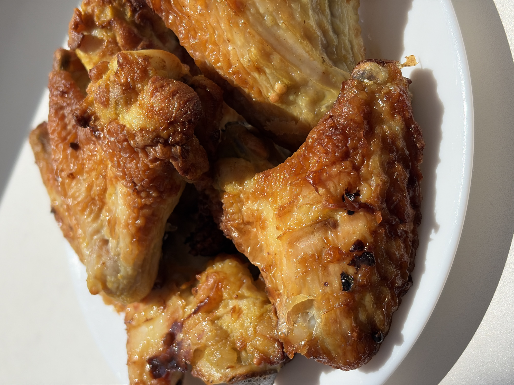 A close-up, overhead shot shows a plate of golden-brown, crispy chicken wings. The wings are arranged somewhat haphazardly on a white, round plate, with some overlapping. The lighting creates strong shadows, highlighting the texture of the cooked chicken skin. The background is a plain, light-colored surface. There are no people or any other identifiable objects in the scene. The location is Unknown, Unknown.