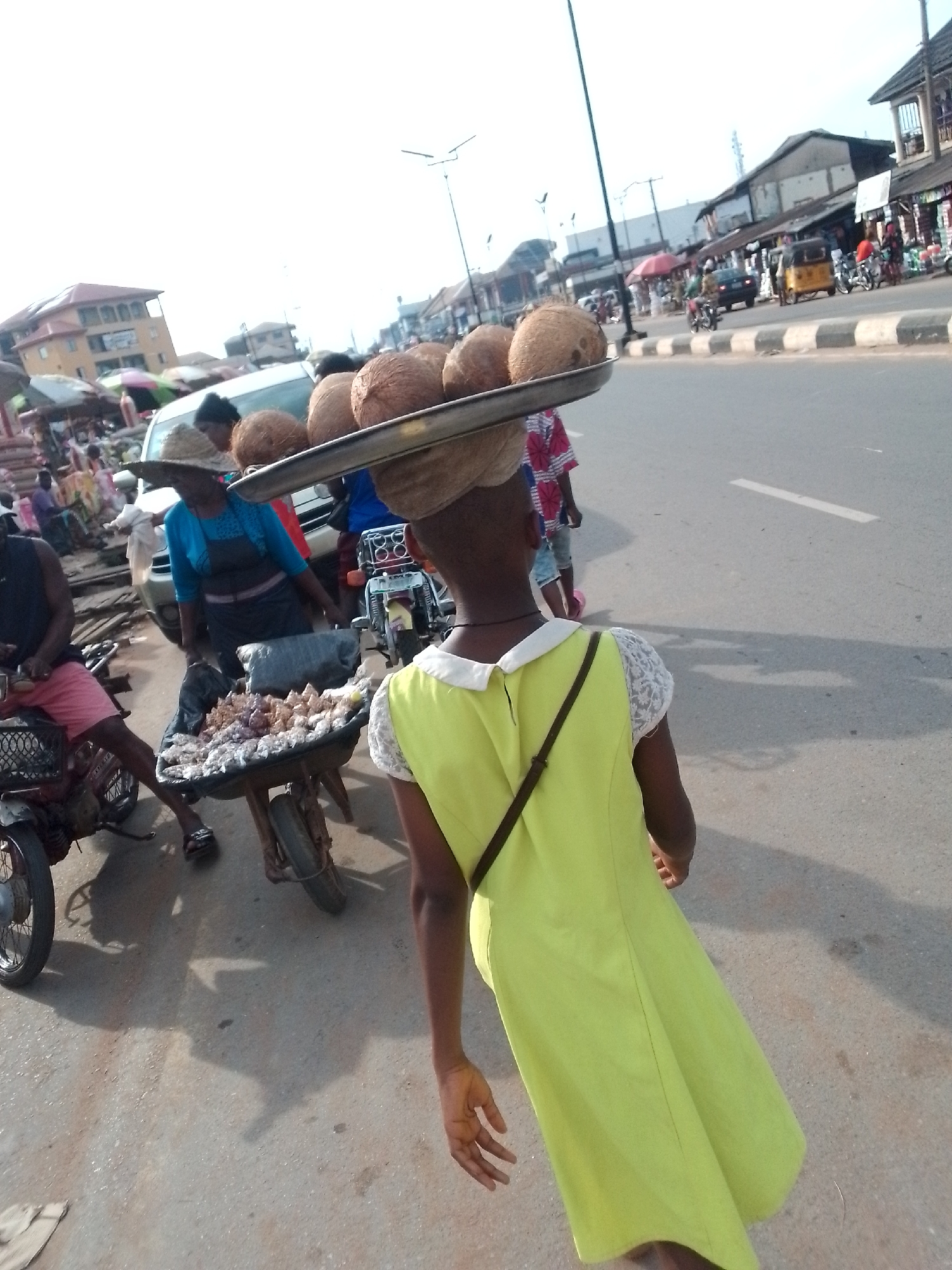 A street scene in Orlu, Nigeria, depicts a young girl carrying a tray of coconuts on her head. The girl, wearing a light green dress with white lace sleeves and a black shoulder strap, walks on the side of a road. A woman wearing a large straw hat stands beside a cart filled with goods, possibly food items. A man in a black shirt and pink shorts sits on a motorcycle next to the cart. Other people, vehicles, and buildings line the street, which has a white line marking the lanes. Street vendors appear to be selling goods. The setting suggests an active commercial area.
