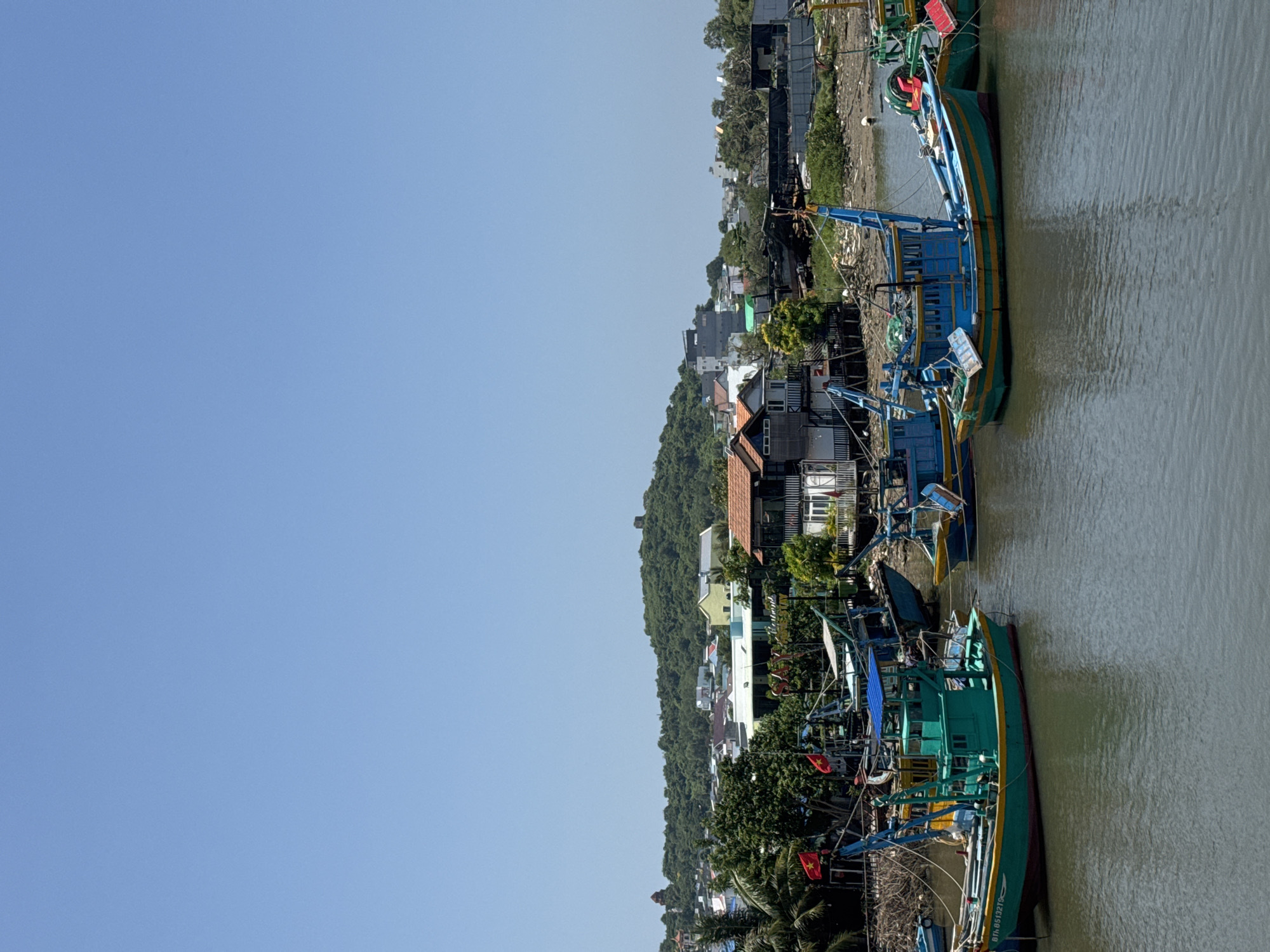 This is a photograph taken in the city of Phu Thuy, Vietnam, on a clear day. The scene depicts a body of water with several fishing boats docked along the shore. The boats are colorful, with green, blue, and yellow being prominent colors. Some of the boats have flags, including Vietnamese flags. In the background, there are houses and buildings with red tile roofs, interspersed with lush green trees and vegetation. A gently sloping, tree-covered hill rises in the distance, with some structures visible on its slopes and summit. The water appears murky, and the shoreline shows evidence of sediment and debris. The sky is a clear, bright blue. There are no people visible in the immediate foreground of the photograph, but the presence of the boats suggests a fishing community. The overall impression is a working harbor in a coastal or riverine town.