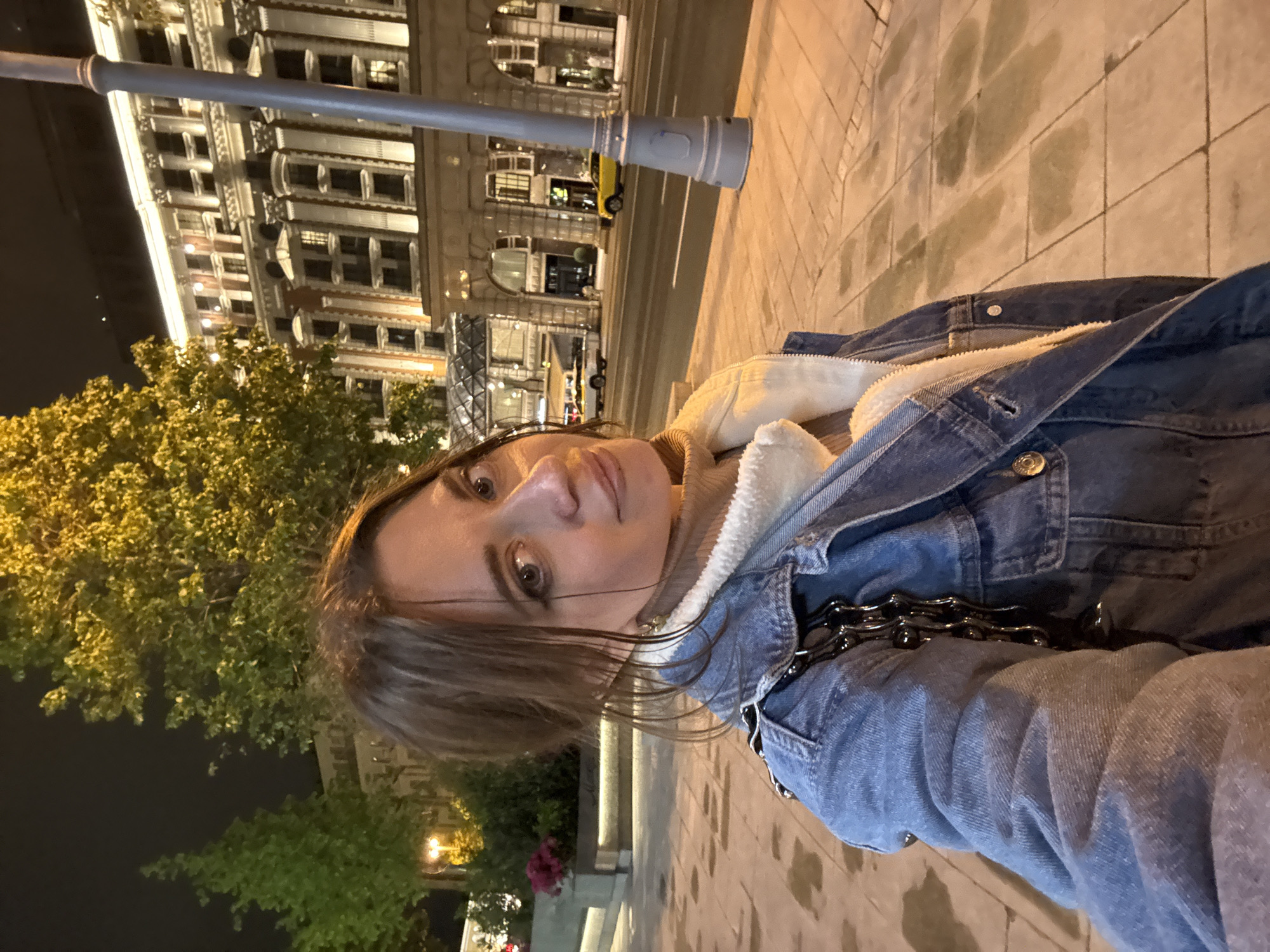A medium-shot perspective shows a fair-skinned woman with brown hair posing in front of an indistinct building on a city street at night. The woman is wearing a blue denim jacket with a white, shearling-like lining. A dark, metallic chain-like strap is visible on her shoulder. The street appears to be paved with light-colored tiles. A streetlight and trees are visible in the background. Based on the architectural style and ambiance, the location appears to be Moscow, Russia.
