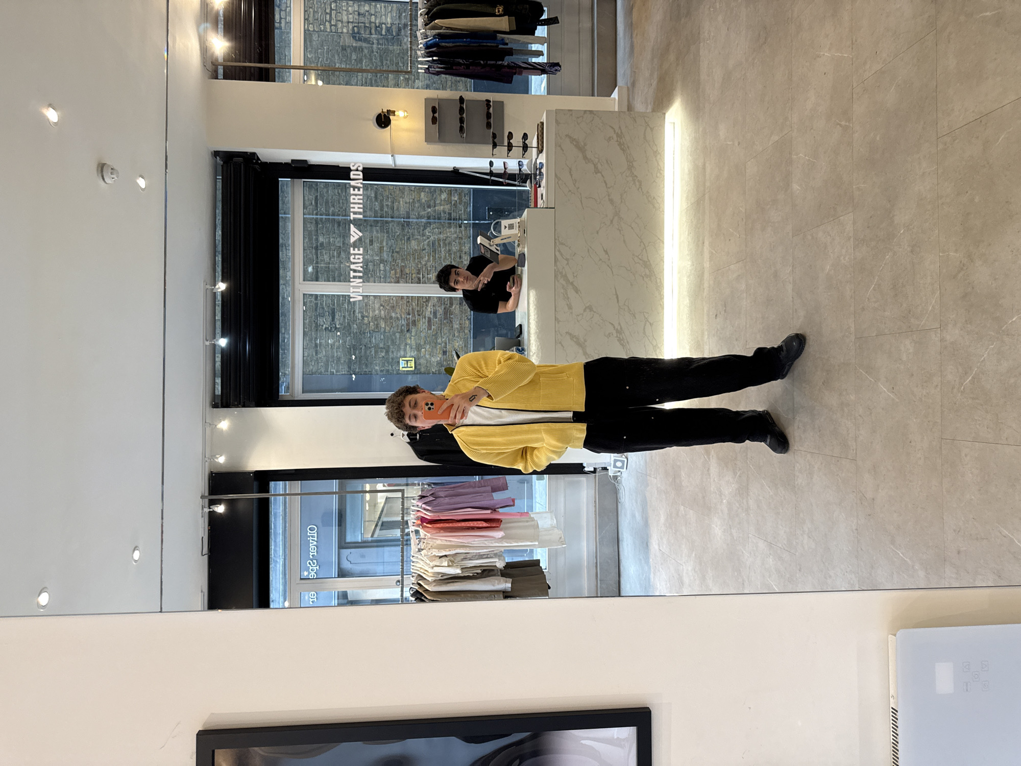 A mirror selfie is being taken inside a modern retail store, likely a clothing or vintage shop in London, United Kingdom. The primary subject is a person holding a smartphone to take the photo, captured from the chest down in the mirror's reflection. This person is wearing a bright yellow ribbed jacket over a white top, black trousers, and dark shoes.
The store's interior is brightly lit, suggesting daytime. It features light beige tiled flooring and white walls, creating a clean and contemporary atmosphere. In the background of the reflection, behind the selfie-taker, clothing racks display a variety of garments in shades of pink, purple, white, and olive green.
To the right, a white counter with a gray marble-like pattern holds sunglasses and other accessories. Behind this counter, another man wearing a black t-shirt is visible, leaning forward with his left arm resting on the counter and his right hand making a gesture. He appears to be interacting with the person taking the photo or observing the scene. A cash register or point-of-sale system is also visible on the counter.
The store's name, "VINTAGE THREADS," is prominently displayed in reverse, reflected on a large window or sign in the background. Through another window further back, the reflection of an external brick building can be seen. Various light fixtures, including recessed ceiling lights and decorative wall lights, illuminate the space. A white fire alarm or smoke detector is mounted on the wall to the left of the main scene. In the bottom-right corner, part of a white electronic device with a digital display is visible. The overall setting is well-maintained and stylish.