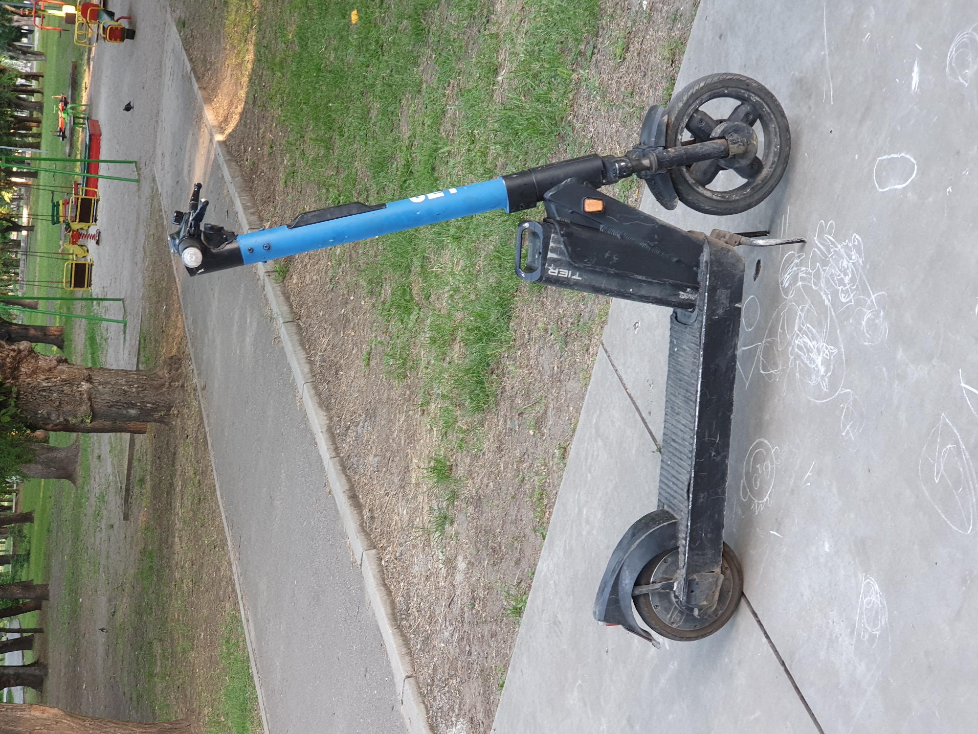 An electric scooter, branded "TIER," is parked on a concrete sidewalk in front of a grassy area with trees and a playground in the background. The scooter is black and blue, with a black stem and handlebars. The front wheel is black with silver spokes, and the rear wheel is also black. Chalk drawings of various figures are visible on the sidewalk around the scooter. In the background, there are swings, a sandbox, and a seesaw. Several birds are visible in the grassy area and on the playground. The sun appears to be shining, casting shadows.