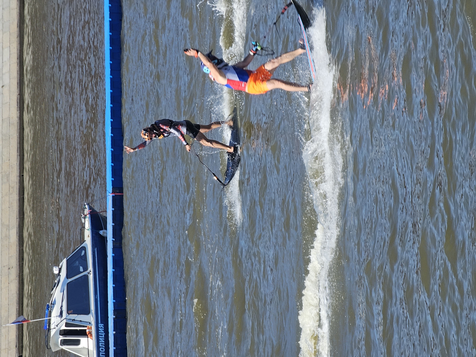 A police boat is moored to a blue floating barrier on a body of water. Two individuals on personal watercraft are in the water. One individual, in the foreground, is wearing orange shorts and a red, white, and blue top, and is on a black watercraft with a blue and red stripe. They are holding a black pole with a cord attached. The second individual, further back and to the left, is wearing a patterned top and black shorts, and is on a black watercraft. They are also holding a black pole with a cord. The water is choppy, with whitecaps visible. The sky is blue and the sun is shining. The background shows a concrete embankment.