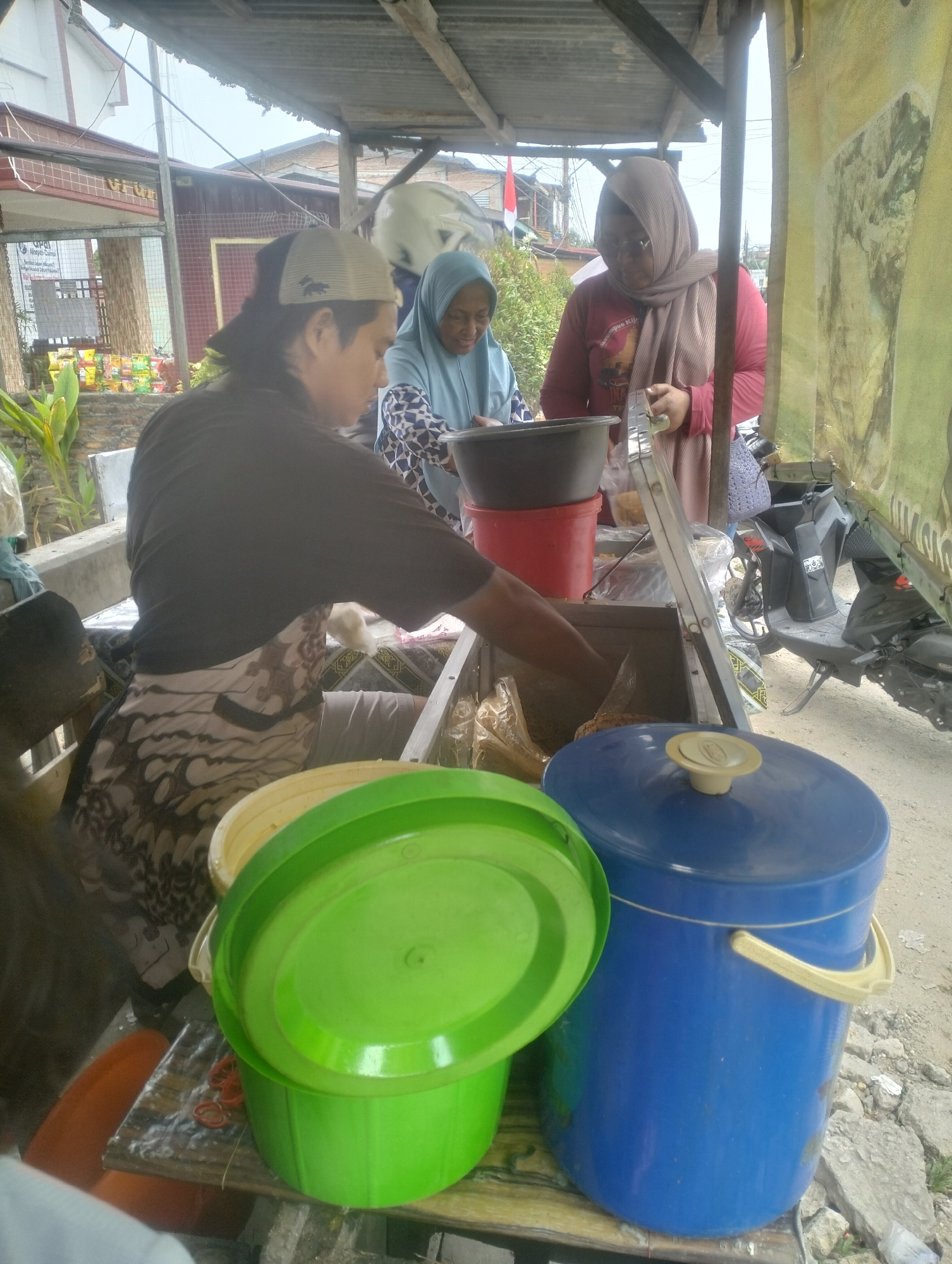In Dumai City, Indonesia, an outdoor food stall is pictured. Three individuals are present. On the left, a person wearing a dark shirt and a patterned apron is attending to food preparation. In the center, a woman in a blue hijab and patterned top is seen. To the right, another woman in a beige hijab and a pink long-sleeved shirt appears to be interacting with a customer or colleague. The stall is covered by a corrugated metal roof supported by wooden posts. Various containers, including large blue and green buckets, are visible, along with a red bucket and a black basin. A motorcycle is parked behind the stall. The ground is dirt and rocks.