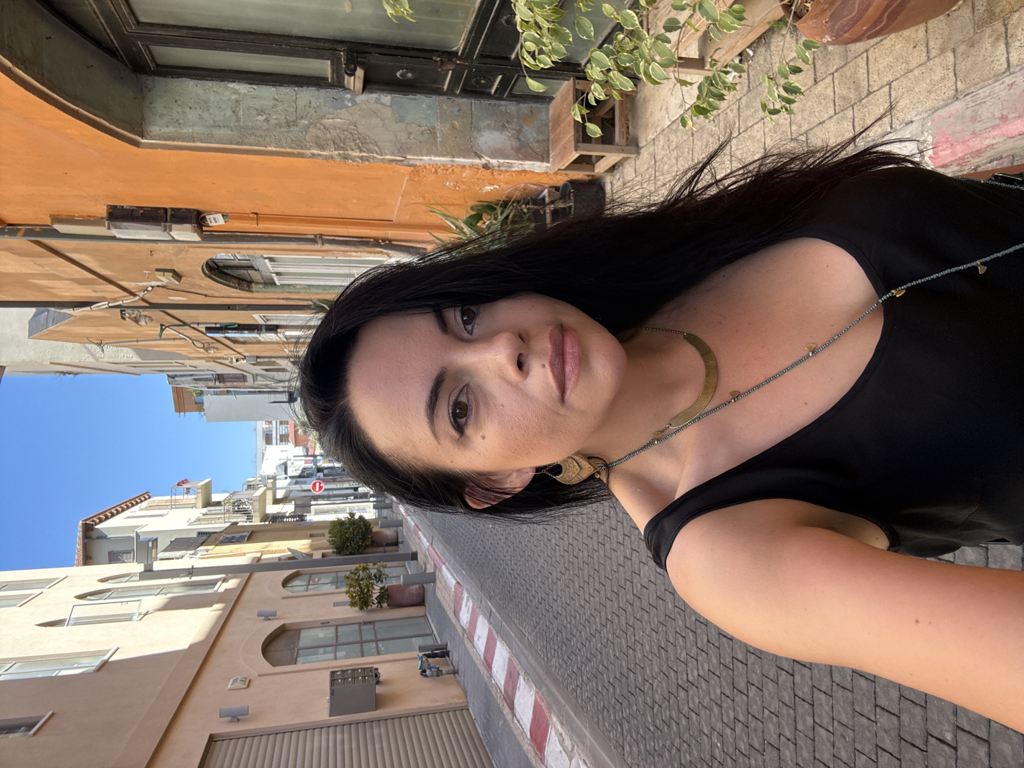 A woman with dark hair and brown eyes takes a selfie in a narrow, sunlit street in Tel Aviv, Israel. She is wearing a black sleeveless top and a multi-layered necklace with a curved pendant and small blue beads. Her left side is visible, showing her shoulder and arm. She has large, gold, drop earrings.
The street is paved with grey cobblestones and bordered by a sidewalk with red and white striped markings. The buildings on either side are narrow and tall, with a mix of architectural styles. The building on the left is a pale peach color with arched windows and a grey garage door. The building on the right is a vibrant orange with a textured facade and a dark wooden door. Potted plants, including a ficus, are visible in front of the orange building. The sky above is a clear blue. The scene appears to be during the daytime.