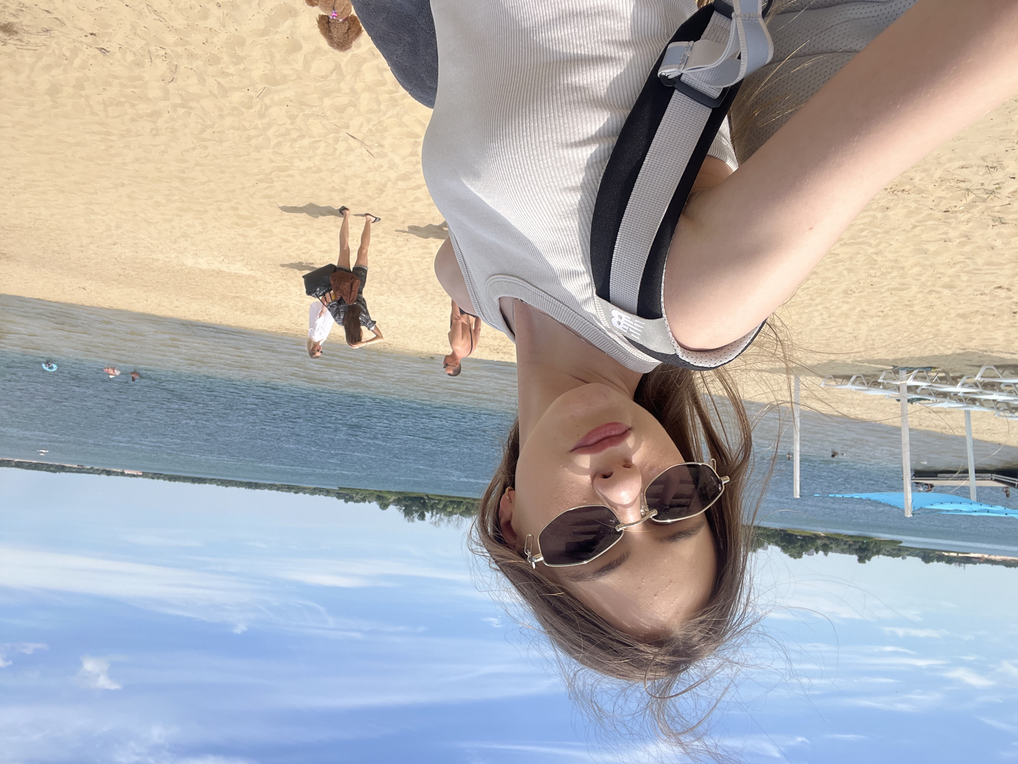 A young woman with long, brown hair and sunglasses is taking a selfie on a sandy beach. She is wearing a light-colored, ribbed tank top and a backpack. In the background, a body of water is visible, with a few people swimming and a dock with a blue canopy. The sky is blue with scattered clouds. The beach is lined with lounge chairs. The location is identified as Yelyzavetivka, Ukraine.
