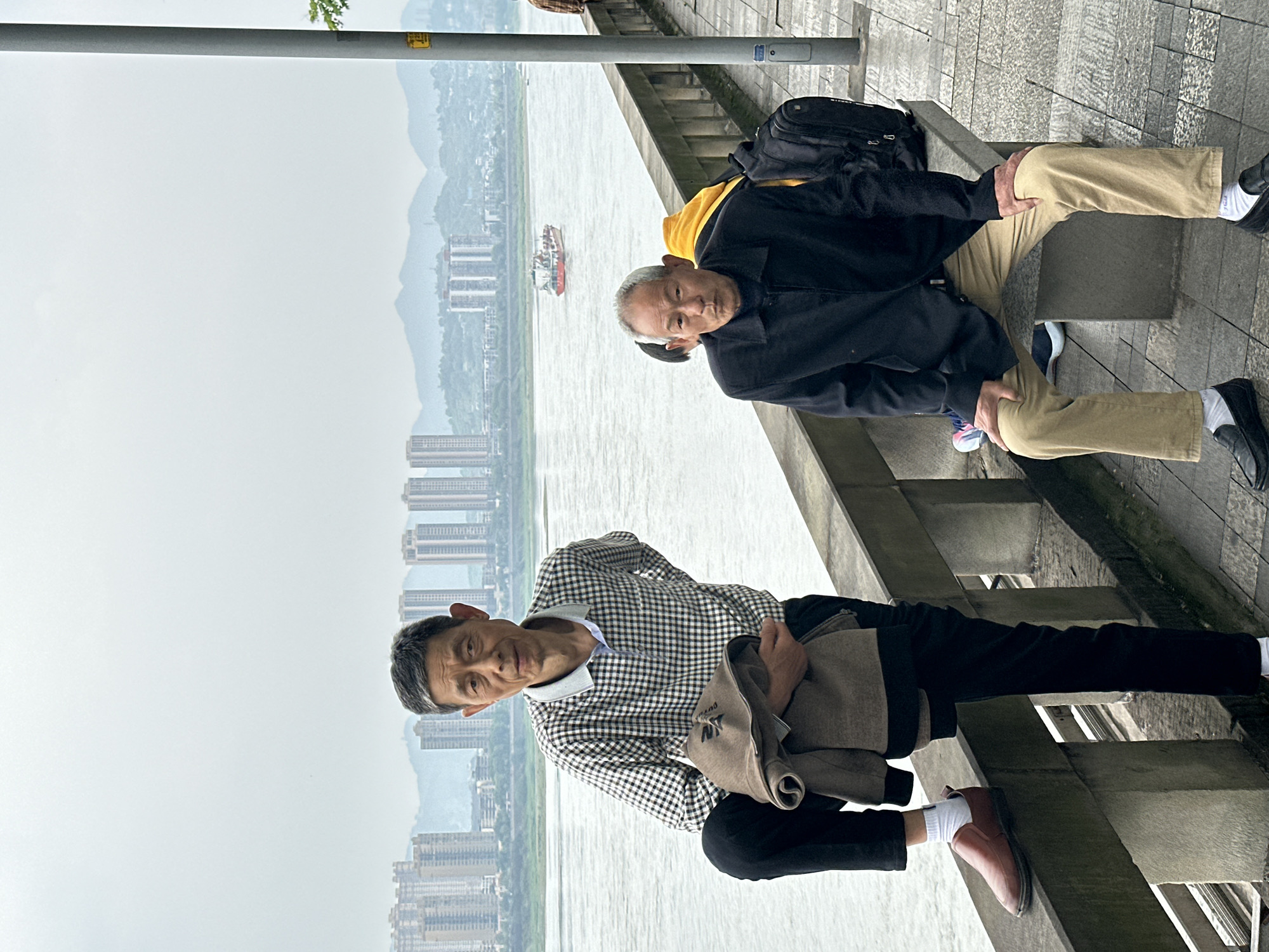 Two men are seated on a concrete structure beside a body of water. Behind them is a cityscape of high-rise buildings in Jiangjin, China. A boat is visible on the water. The man in the foreground wears a checkered shirt and black pants, while the man in the background wears a black jacket over a yellow shirt, with khaki pants. The men are facing the camera.
