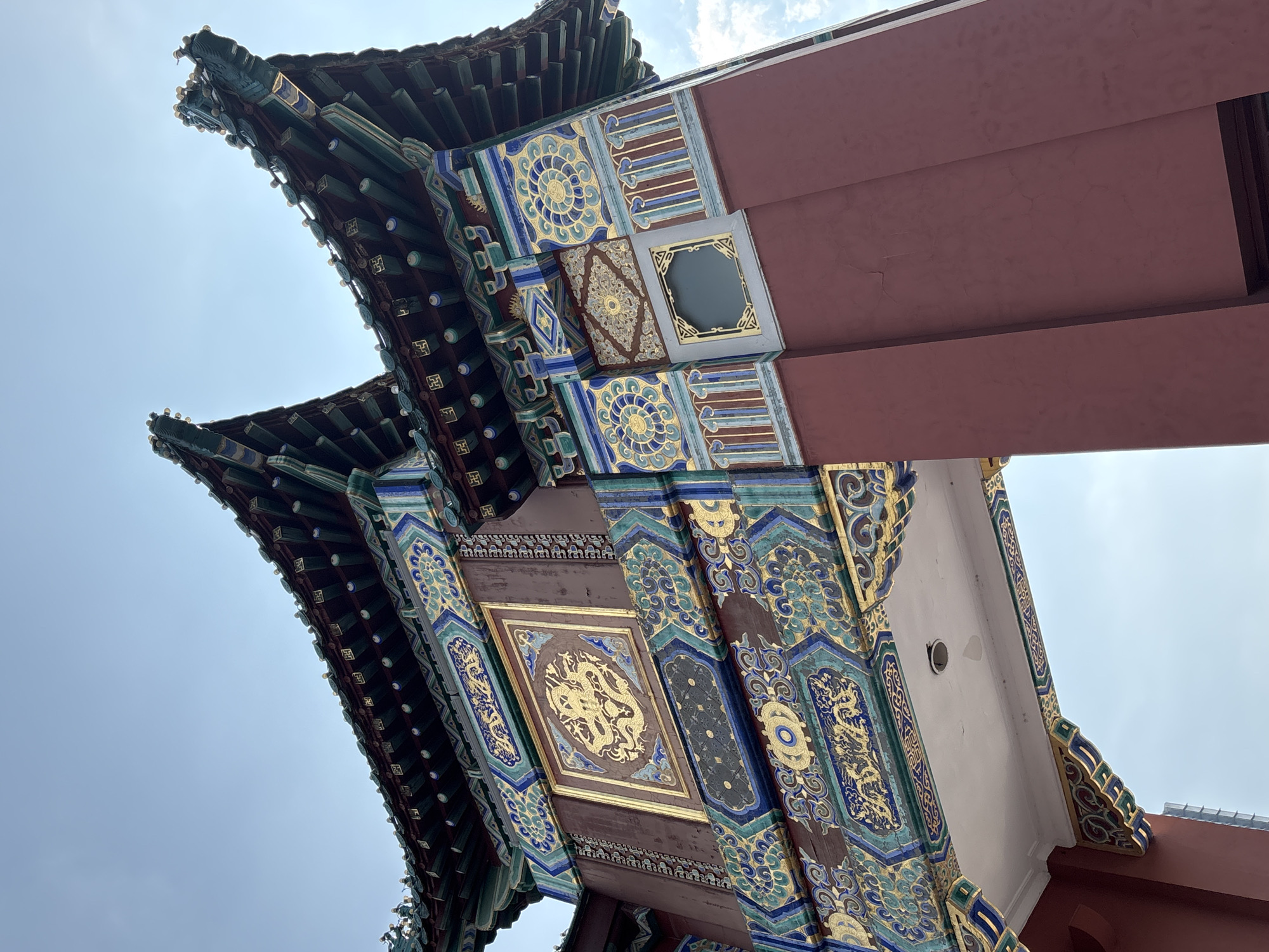 The image shows a close-up, low-angle view of a richly decorated traditional Chinese archway, likely a pailou. The archway features a multi-tiered, upturned roof covered in dark green tiles with ornate, colorful decorations along the eaves. The body of the archway is painted a deep red, adorned with intricate gold and blue patterns, including a prominent golden dragon motif within a square panel. Sections of the archway showcase geometric designs and floral patterns in vibrant blues, greens, and golds. The sky above is a clear, pale blue with some light clouds visible. The image appears to have been taken during the day. The context provided indicates the location is Yuzhong District, China, suggesting this is a cultural landmark within that area. There are no people or overt actions depicted; the focus is solely on the architectural elements.