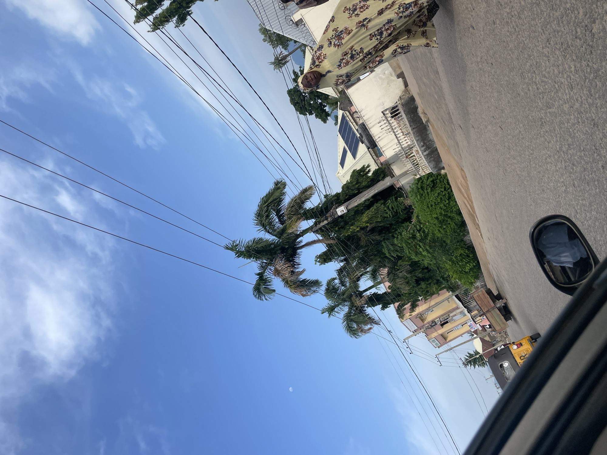 The image depicts a street scene in Jalingo, Nigeria. A woman wearing a yellow and floral patterned hijab and dress is walking on the right side of the road. In the background, several palm trees and other lush green vegetation are visible. Buildings with solar panels on their roofs and decorative metal fences line the street. Overhead, a network of power lines crisscrosses the sky. A yellow auto-rickshaw, a common mode of transport in Nigeria, is seen driving on the road, along with a flatbed truck. The sky is a clear blue with scattered white clouds, and the moon is faintly visible. The perspective of the photograph suggests it was taken from inside a moving vehicle, with a portion of the car's interior, including a side mirror reflecting the scene, visible in the foreground. The overall impression is of a typical day in a Nigerian city.