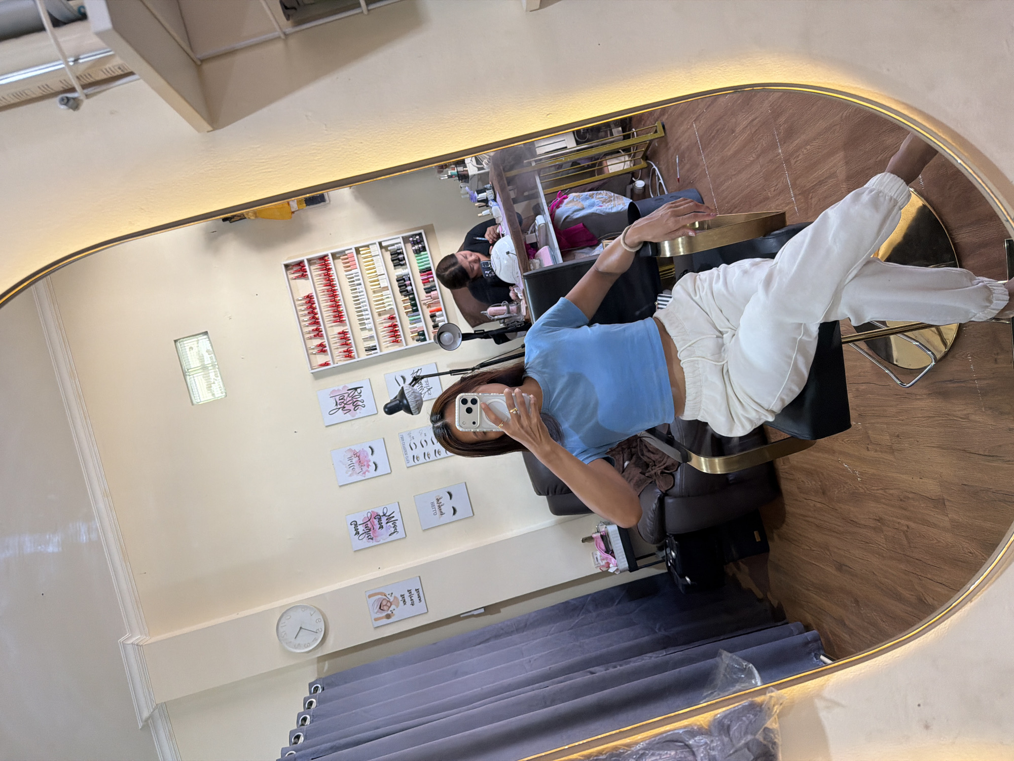 The image displays a mirror selfie taken inside a beauty salon in North Kuta, Indonesia. The main subject is a woman, reflected in a large, horizontally oriented mirror with a golden frame. She is wearing a light blue crop top and white jogging pants, holding a smartphone with a prominent magnetic ring accessory to capture the photo. Her right arm rests on a golden armrest of a salon chair.
In the background reflection, another individual, likely a salon professional, is partially visible, seemingly engaged in a beauty service such as a manicure or pedicure. The salon walls feature organized white shelving units stocked with numerous bottles of nail polish in various colors, including red, white, gold, and black. Other salon equipment, such as a task lamp and product trolleys, are also reflected.
Decorative signs adorn the light-colored walls, displaying beauty-themed text and graphics. Visible phrases include "Boss Babe," "Hello Gorgeous," "Life is too short to have boring nails," "Wake up and makeup," and a partially visible "Beauty begins the moment you decide to be yourself." A white circular wall clock is also present. Gray curtains hang along the lower part of the wall, and the floor reflected in the mirror appears to be made of wood. The salon appears well-lit and orderly, creating a professional and inviting atmosphere.