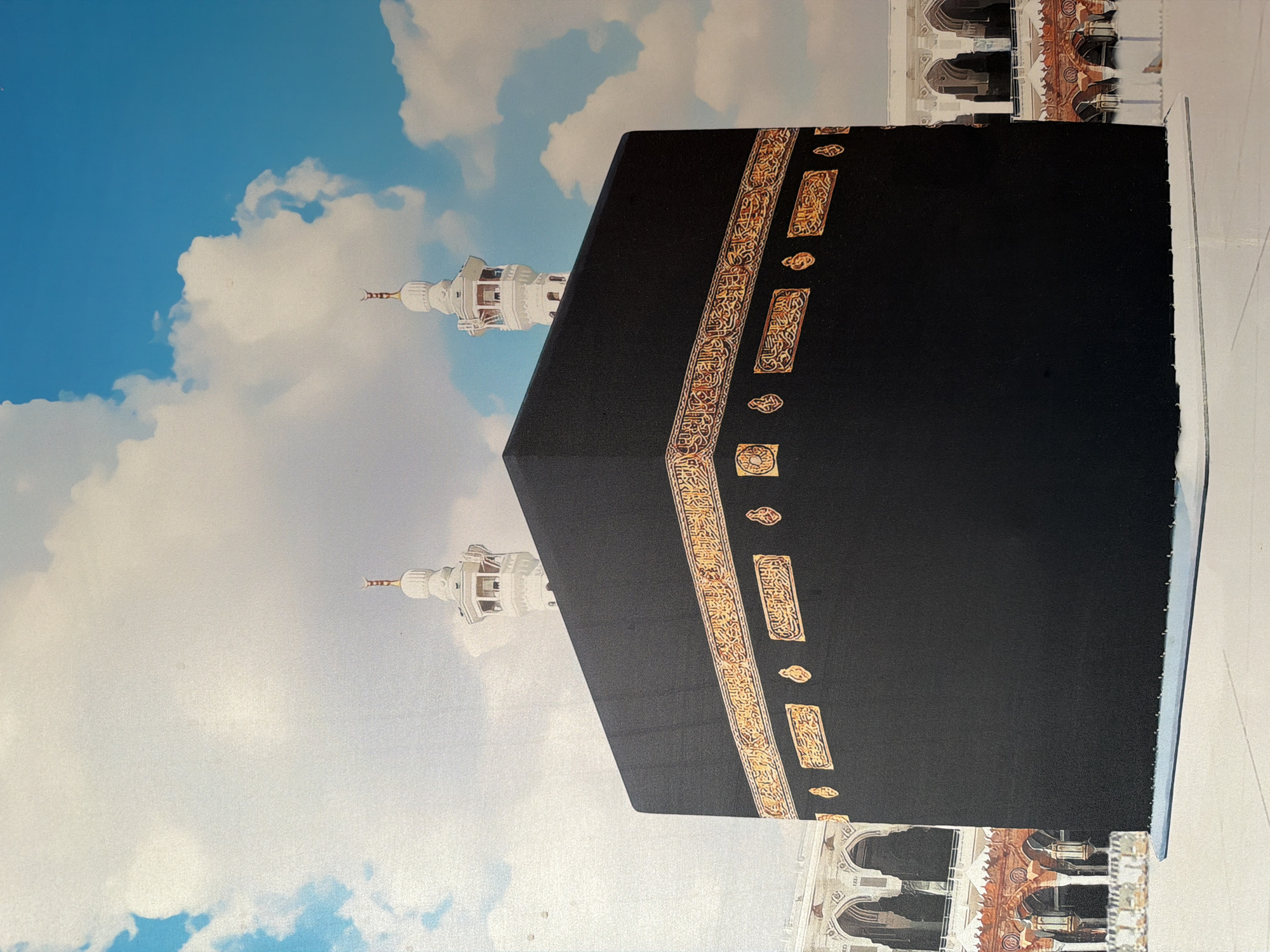 This media file features a vibrant depiction of the Kaaba, the holy site in Mecca, Saudi Arabia, likely a print or poster, photographed from Akkuzovo, Russia. The image is oriented vertically, showing the Kaaba prominently positioned on the right side, set against a bright blue sky with fluffy white clouds on the left.

The Kaaba itself is a large, cubic structure, draped in its traditional black covering (Kiswa). This covering is intricately adorned with bands of rich gold-embroidered Arabic calligraphy, which stand out against the dark fabric. The calligraphy appears in vertical strips and smaller ornate motifs, adding significant detail and cultural context to the main subject.

In the background, on the left side, two elegant, light-colored minarets are visible, rising into the sky from behind the Kaaba. Their architectural details, including small balconies and pointed tops, are discernible. The sky behind them is a clear, inviting blue with scattered white clouds, suggesting daytime.

Towards the bottom right of the image, parts of other architectural structures can be seen, characterized by arched windows and ornate, reddish-brown details, indicative of the surrounding Grand Mosque complex. No people or specific activities are visible, presenting a serene and monumental view of the sacred site. The image emphasizes the architectural grandeur and symbolic importance of the Kaaba and its immediate surroundings.