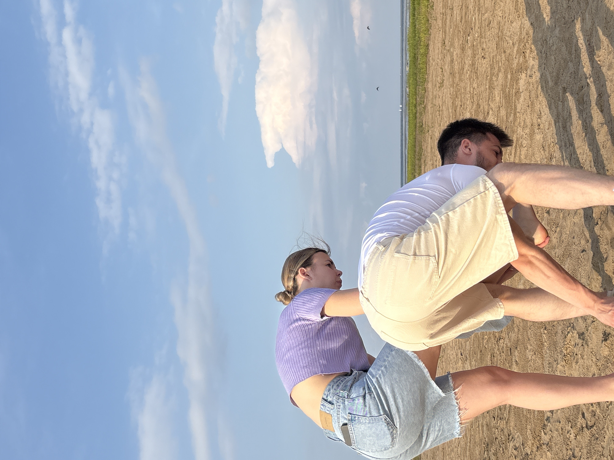 Two individuals are seen on a sandy beach in Koshkino, Russia, bending down and appearing to search for something on the ground. The person on the left, identified as female, wears a purple cropped shirt and light-wash denim shorts. The person on the right, identified as male, is dressed in a white t-shirt and beige shorts. The background features a calm body of water with distant sailboats and green vegetation along the shoreline under a bright blue sky with scattered white clouds. The overall environment suggests a tranquil, outdoor setting, possibly during daytime.