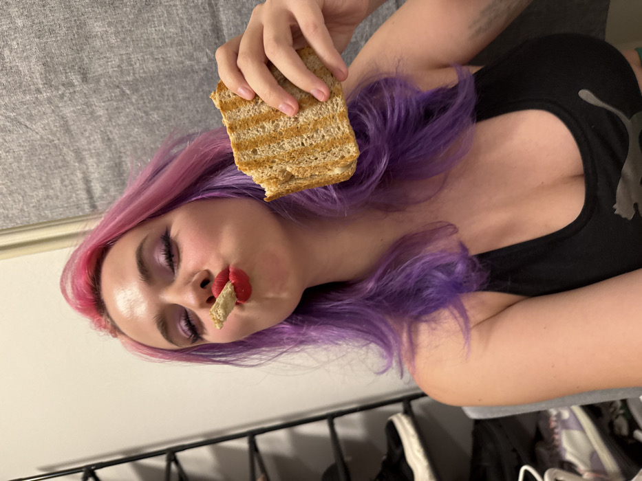 A woman with pink and purple hair, dark eyeshadow, and bright red lipstick, holds a peanut butter sandwich over her head and another piece of bread in her mouth. She is lying down, wearing a black tank top. A grey blanket is in the background, and a clothing rack is visible at the bottom. The image is rotated so the bottom of the frame is on the left.