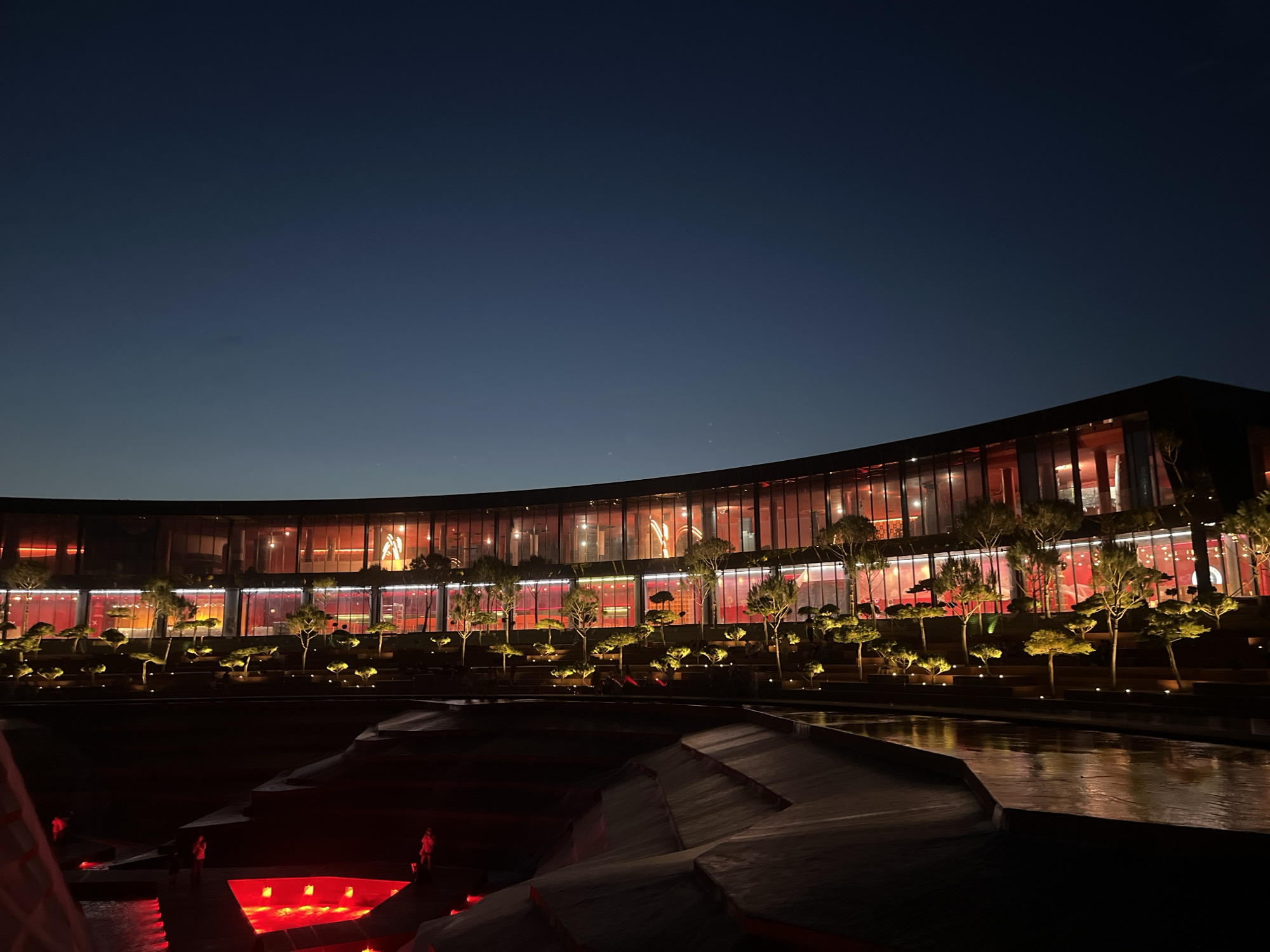The image depicts a modern, curved building with extensive glass windows, illuminated by red and orange lights from within. The building is set against a dark, twilight sky. In front of the building, a tiered landscape features rows of illuminated, manicured trees. Below the trees, a dark water feature or pool reflects the lights. Several figures, appearing as silhouettes, are visible in the foreground, some standing near water illuminated by red lights, possibly indicating a decorative water feature or pool. The overall impression is one of contemporary architecture and landscape design, highlighted by artificial lighting at dusk. The location is identified as the Monument of Memory and Glory in Krasnodar, Russia, which features a unique amphitheater design and reflective pools.
