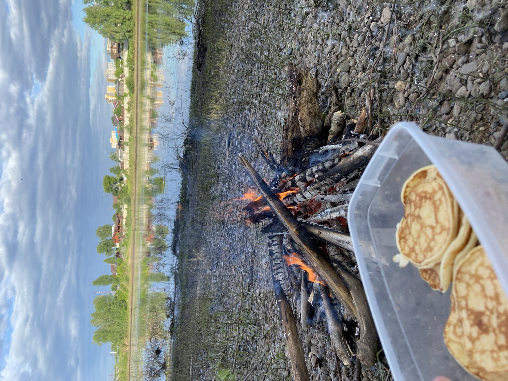 The image presents a riverside scene in the city of Chaikovsky, Russia. In the bottom left foreground, a transparent plastic container holds multiple golden-brown pancakes or blini, partially obscured by a finger. Adjacent to this, a bonfire burns on a rocky riverbank, characterized by small pebbles, larger stones, and sparse green vegetation. Flames and smoke are visible from the burning logs and branches. The midground features a calm body of water, a river, which reflects the sky and structures on the distant shore. The far bank across the river is lined with deciduous trees. Behind these trees, a mixed urban landscape is visible, comprising several single-family houses with sloped roofs and multiple multi-story apartment buildings with light-colored exteriors. The sky above is partly cloudy, displaying patches of blue sky interspersed with white and grey cloud formations. No people are discernible in the scene.