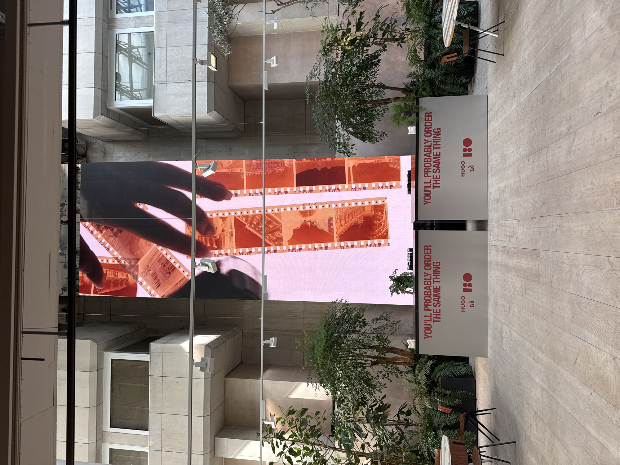 Here is a detailed description of the image:
**Scene:**
The image captures an indoor space in what appears to be a modern building or atrium. The scene is viewed through a window or glass panel, with elements of both the exterior and interior visible.
**Objects:**
* **Large Digital Display:** A sizable digital screen dominates the view, showcasing what seems to be a negative film strip with images. A hand appears to be interacting with the film on the display.
* **Signage:** Beside the display screen, two signs with the inscription "YOU'LL PROBABLY ORDER THE SAME THING" are seen along with "HUGO 180".
* **Vegetation:** The space is adorned with plants, including trees, adding a touch of nature to the modern setting.
* **Furniture:** The scene incorporates tables and chairs arranged for seating.
* **Building Architecture:** The architectural details of the building, including windows and building materials, are visible in the background and as reflections in the glass.
**People:**
No people are directly visible in the scene, although the hand on the display screen indicates human interaction with the technology.
**Environment:**
The setting is a well-lit and contemporary space. The location is speculated to be in the city of London, United Kingdom due to the architectural style and branding. The scene seems to be a combination of retail or display space with architectural elements.