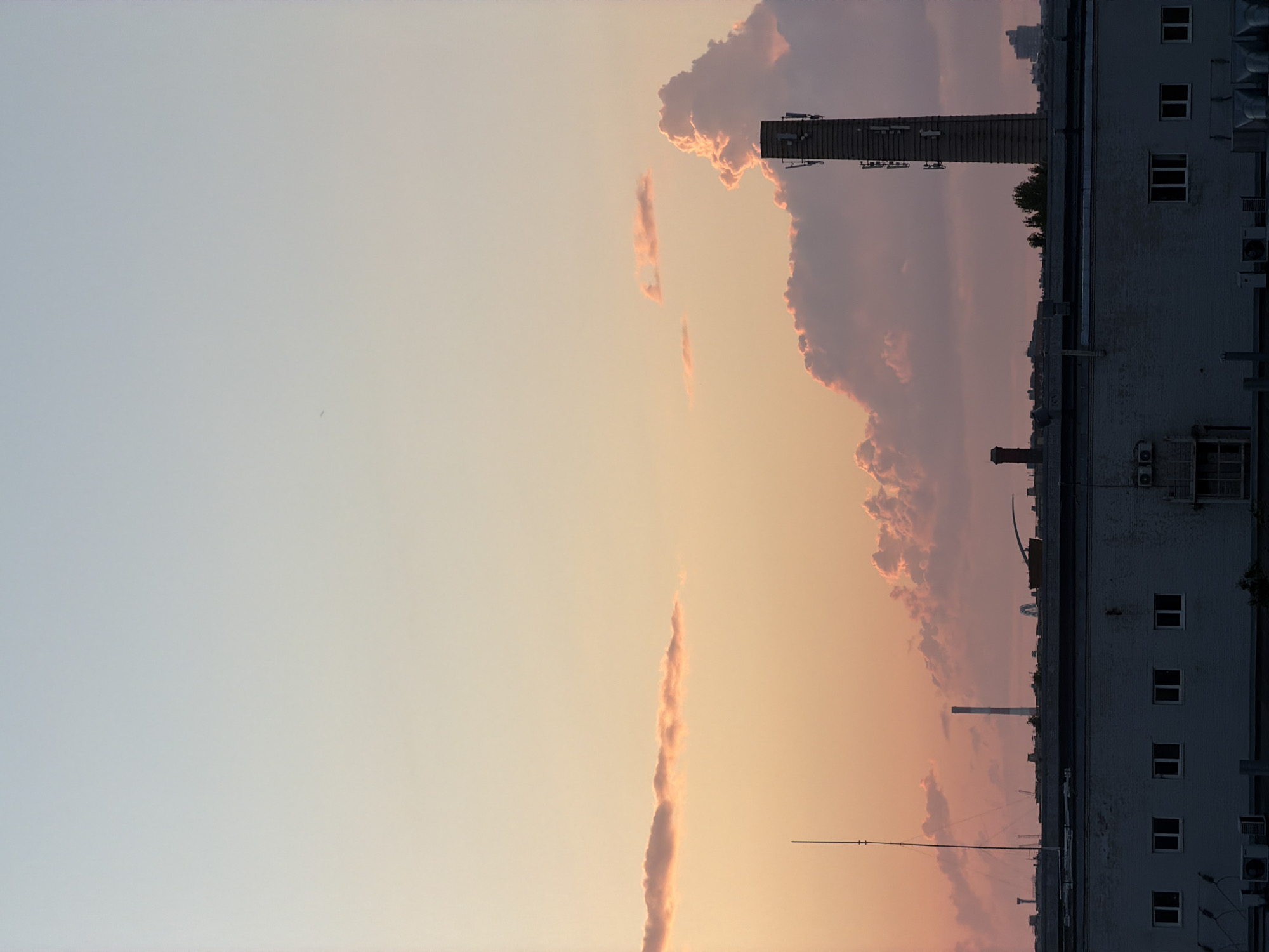 This is a photograph of a cityscape at sunset in Saint Petersburg, Russia. In the foreground, a low-rise industrial building with white brick siding and multiple windows is visible. Above this building, the silhouette of a denser urban landscape stretches across the horizon, with various industrial structures, including chimneys and a cell tower, punctuating the skyline. The sky above is a gradient of soft pastel colors, transitioning from a pale blue at the top to a warm orange and pink near the horizon, with scattered clouds catching the last light of the day. There are no people or animals visible in the frame.