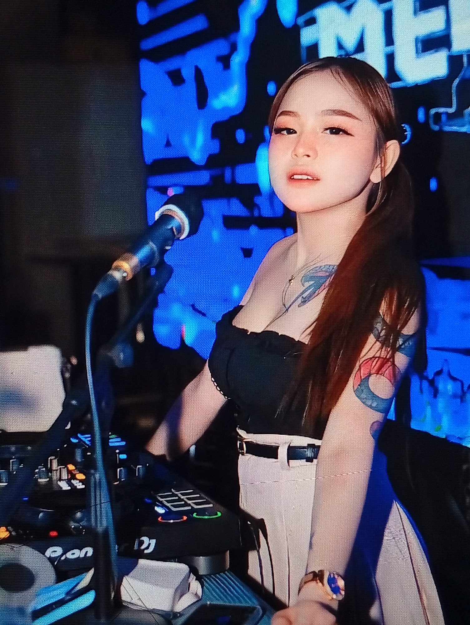 A young woman with brown hair tied in a ponytail, wearing a black off-the-shoulder top and a light-colored skirt, stands behind a DJ console. She has tattoos on her right arm and chest. A microphone is positioned in front of her, and she appears to be looking at the camera. The background features a large screen with blue, abstract graphics. The DJ console has various buttons, knobs, and illuminated displays. The overall setting suggests a nightclub or event venue. The image was taken in Lhokseumawe, Indonesia.