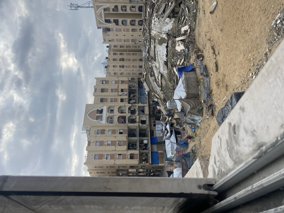 The image presents an elevated view of a heavily damaged urban area in Khan Yunis, Palestinian Territory, under an overcast sky. A concrete window ledge partially frames the bottom-left foreground. The midground features a sandy, debris-strewn ground supporting a cluster of approximately ten makeshift shelters constructed from various tarpaulins in shades of blue, gray, and tan. A single small figure, dressed in red, is observed walking among these temporary structures. To the right of the shelters, a large mound of concrete rubble, twisted rebar, and collapsed building materials is visible. In the background, multiple multi-story residential buildings, light tan with brown window accents, exhibit widespread structural damage. Their facades are pockmarked with numerous impact holes, and many windows are shattered or entirely absent. Lower levels of these buildings show significant collapse, with exposed interiors, some covered by blue tarpaulins. Laundry hangs from a balcony railing on one central building. Communication antennas are discernible on several rooftops.