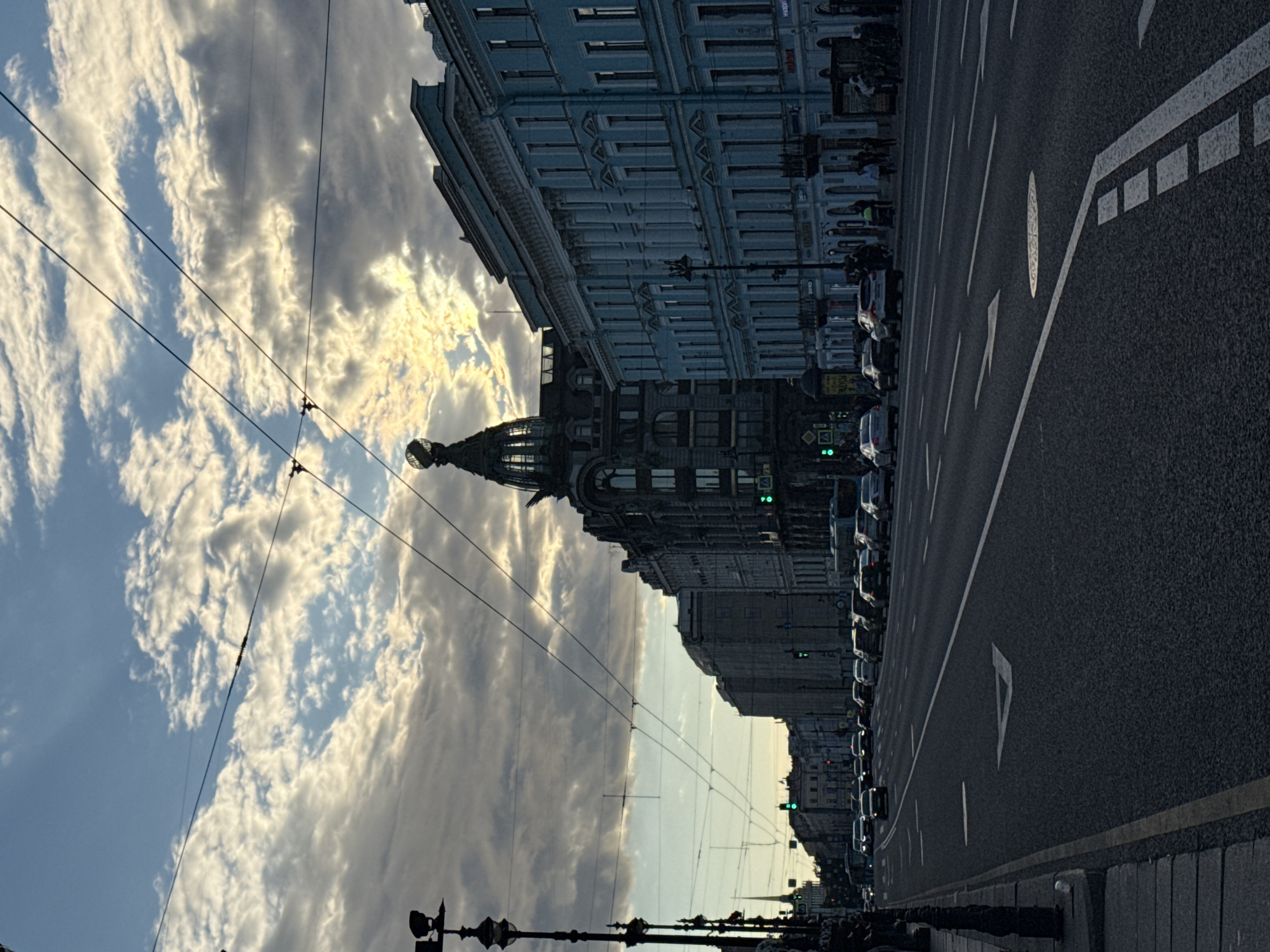 The image shows a street scene in Saint Petersburg, Russia, likely Nevsky Prospekt, as indicated by the architecture and the general layout of a wide avenue with historic buildings and a tram line. The photograph is taken from a low angle, looking down the street towards a cloudy sky. The sky is dramatic, with patches of bright sunlight breaking through dense, textured clouds, creating a luminous effect.
On the right side of the street, a row of ornate, pastel-blue buildings with classical architectural elements like columns and intricate window frames lines the avenue. These buildings appear to be multi-story structures with shops or businesses on the ground floor. Further down the street, a darker, more elaborate building with a distinctive dome-shaped cupola topped with a sphere is visible. This building is set back from the road and stands out due to its unique architectural style.
On the left side of the street, the view is partially obscured by a lamppost and a dark, ornate fence or railing along the sidewalk. More buildings line this side of the street, appearing to be of similar historical style to those on the right. A tall, slender spire of a church or government building can be seen in the distance on the far left.
The street itself is a multi-lane asphalt road with clear lane markings, including directional arrows painted in white. Several cars are visible driving on the road in both directions, some appearing to be in traffic. Traffic lights, indicated by green lights, are visible at intersections further down the street. A few people can be seen walking on the sidewalks of the buildings. The overall impression is one of a bustling city street during a transitional period of the day, possibly late afternoon or early evening, given the dramatic lighting and cloud cover. The scene captures a sense of urban grandeur and the dynamic atmosphere of Saint Petersburg.