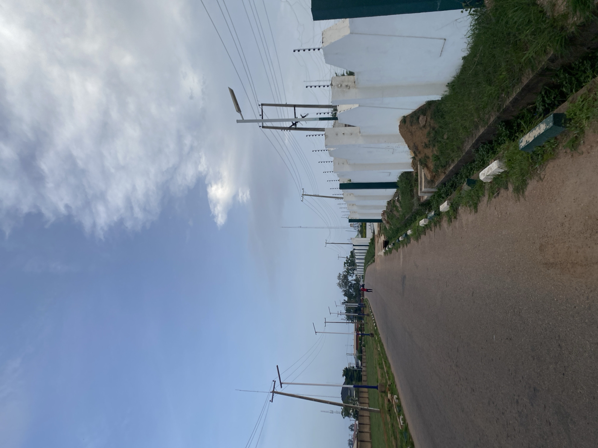 The image shows an asphalt road in Barikin Jirga Summa, Nigeria, lined with a white wall topped with electrical fencing on the right. A drainage ditch runs alongside the road, filled with green grass and vegetation. On the left side of the road, there is a grassy area with several utility poles and streetlights. In the distance, a single person is walking on the road. The sky is partly cloudy with a mix of blue sky and white clouds.