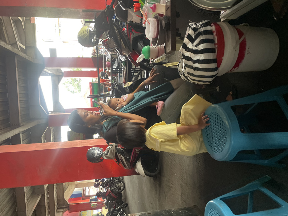The image depicts an outdoor or semi-covered market/food court area in Nganguk, Indonesia. In the foreground, an adult female with dark hair, wearing a dark top and light jeans, is seated while actively using a white smartphone held to her face. An infant is secured in a teal fabric baby carrier on her front, with pink baby shoes visible near its feet. To her left, a young child in a yellow dress sits on a bright blue plastic stool, facing slightly away from the camera.

To the right of the adult, a dark table holds several items: a red-colored beverage in a clear cup with a straw, additional plastic cups and bowls, a white tissue dispenser with a green lid, and a baby bottle. A black and white striped plastic bag rests on top of two stacked white plastic buckets with red rims. In the background, multiple motorcycles and scooters are parked, including a white and red scooter with a black helmet. The area is characterized by thick, square red support pillars and an overhead concrete structure, indicating a public, commercial setting under natural daylight.