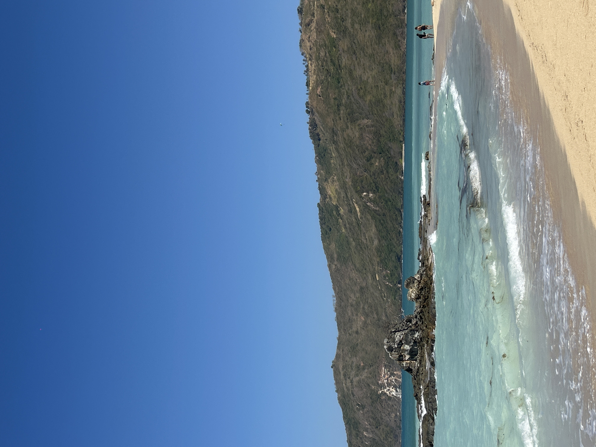 A tropical beach scene unfolds under a clear blue sky. In the foreground, the sandy beach meets the turquoise waters of the ocean. Small waves with white foam lap the shore, carrying bits of seaweed. Further out, several dark rocks protrude from the water, some forming a small island with jagged peaks. In the middle ground, a line of people, two adults and two children, walk along the water's edge. In the background, a lush green mountain range slopes down towards the sea, its slopes covered in dense vegetation. The overall scene evokes a sense of tranquility and natural beauty. The specific location in the city of Kute, Indonesia, is not discernible from the image itself.
