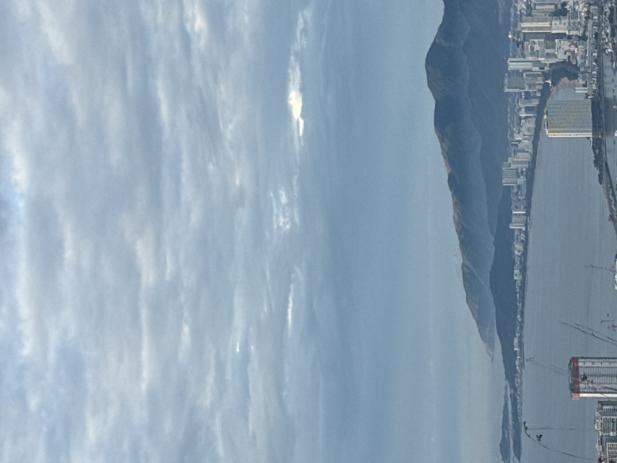 The image shows a view of the city of Bac Nha Trang Ward, Vietnam, along the coast. There is a large mountain range running along the coastline. Buildings and infrastructure line the coast. In the foreground, there is a hazy sky with visible clouds. The image appears to have been rotated.