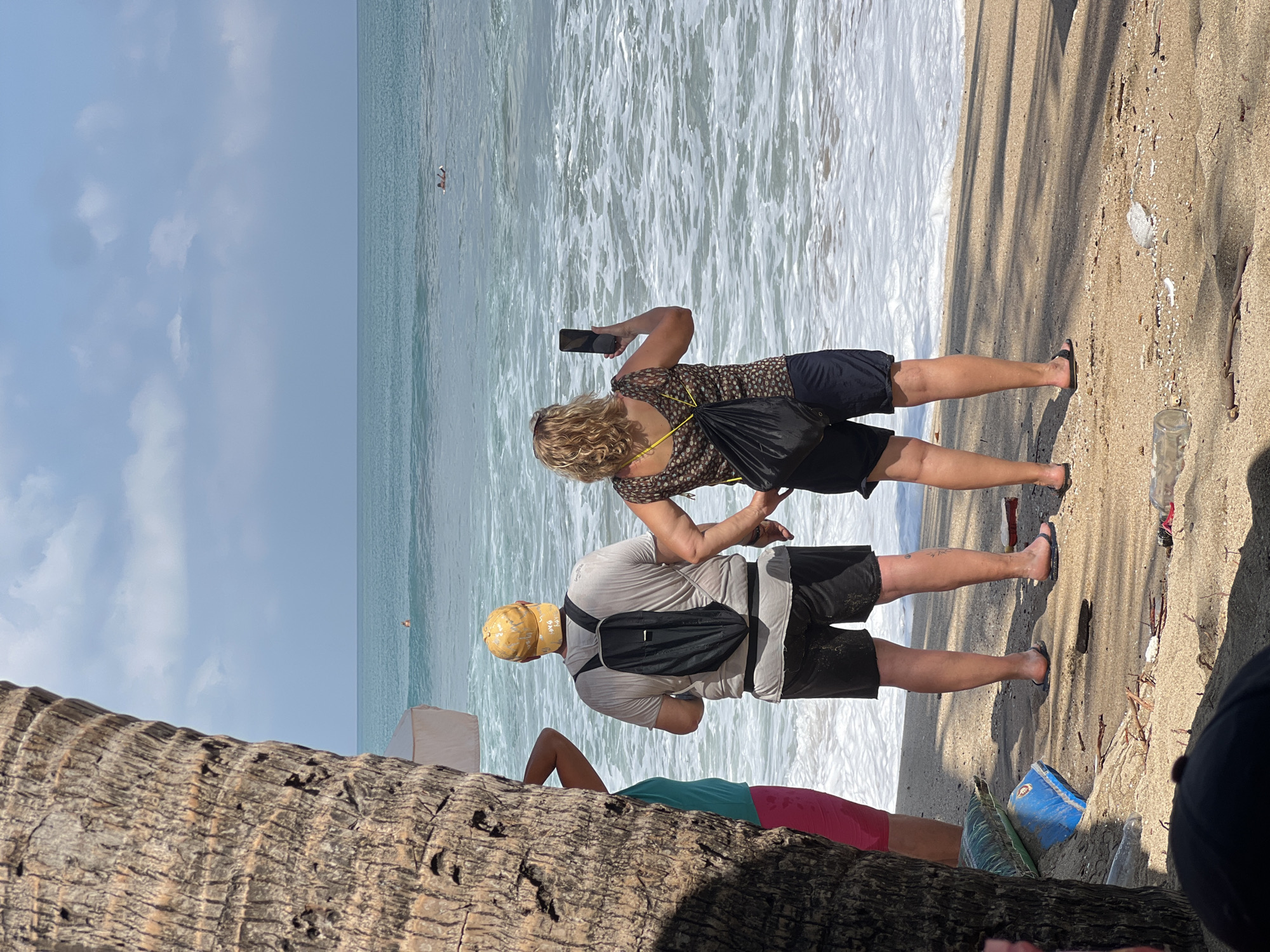 The image depicts a beach scene on a sunny day with some clouds in the sky. In the foreground, a large palm tree trunk is visible on the left. Standing on the sandy beach, facing the ocean, are three people. On the left, partially visible, is a person wearing a turquoise top and pink shorts. In the center, a man wearing a grey t-shirt, black shorts, and a yellow cap has a black backpack on. To the right, a woman with blonde curly hair, wearing a patterned top and dark shorts, is holding up a smartphone to take a photo. She also has a black backpack slung over her shoulder. The ocean is a calm blue with white waves breaking on the shore. Two individuals can be seen swimming or wading in the water further out. The beach is littered with some debris, including plastic bottles and branches. The environment suggests a tropical location, consistent with Kuta, Indonesia.