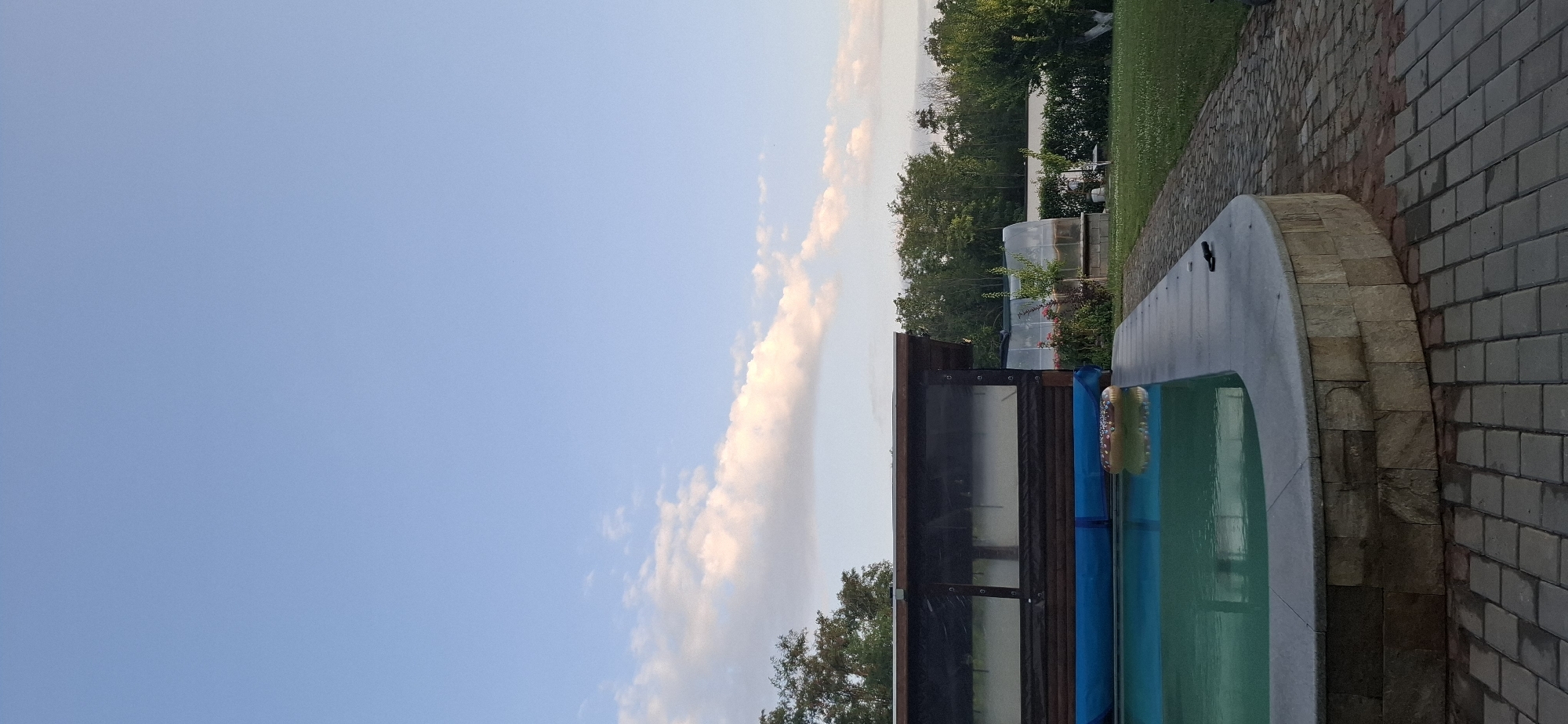 An outdoor swimming pool with a stone surround is visible, with a donut-shaped inflatable float in the water. A wooden structure with transparent coverings is situated next to the pool, and beyond that, a greenhouse and lush green trees are present. A paved walkway lines the edge of the pool area, leading into a grassy yard. The sky above is a clear blue with scattered white clouds, suggesting it is daytime. The scene appears to be in a residential backyard.