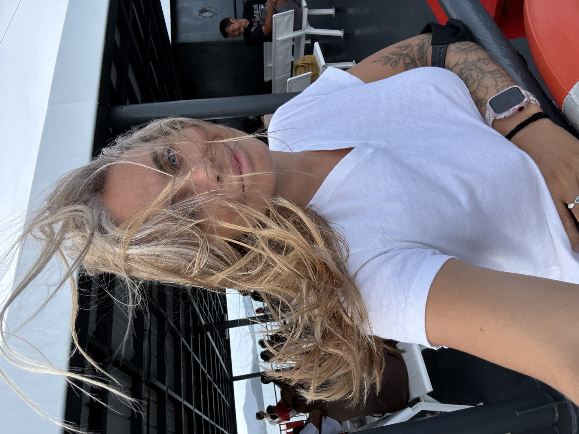 The image shows a woman on a ferry with her hair blowing in the wind. She is wearing a white t-shirt and has tattoos on her arm. In the background, there are other passengers and the ocean. The location is likely a ferry in the waters off Ko Samui District, Thailand.