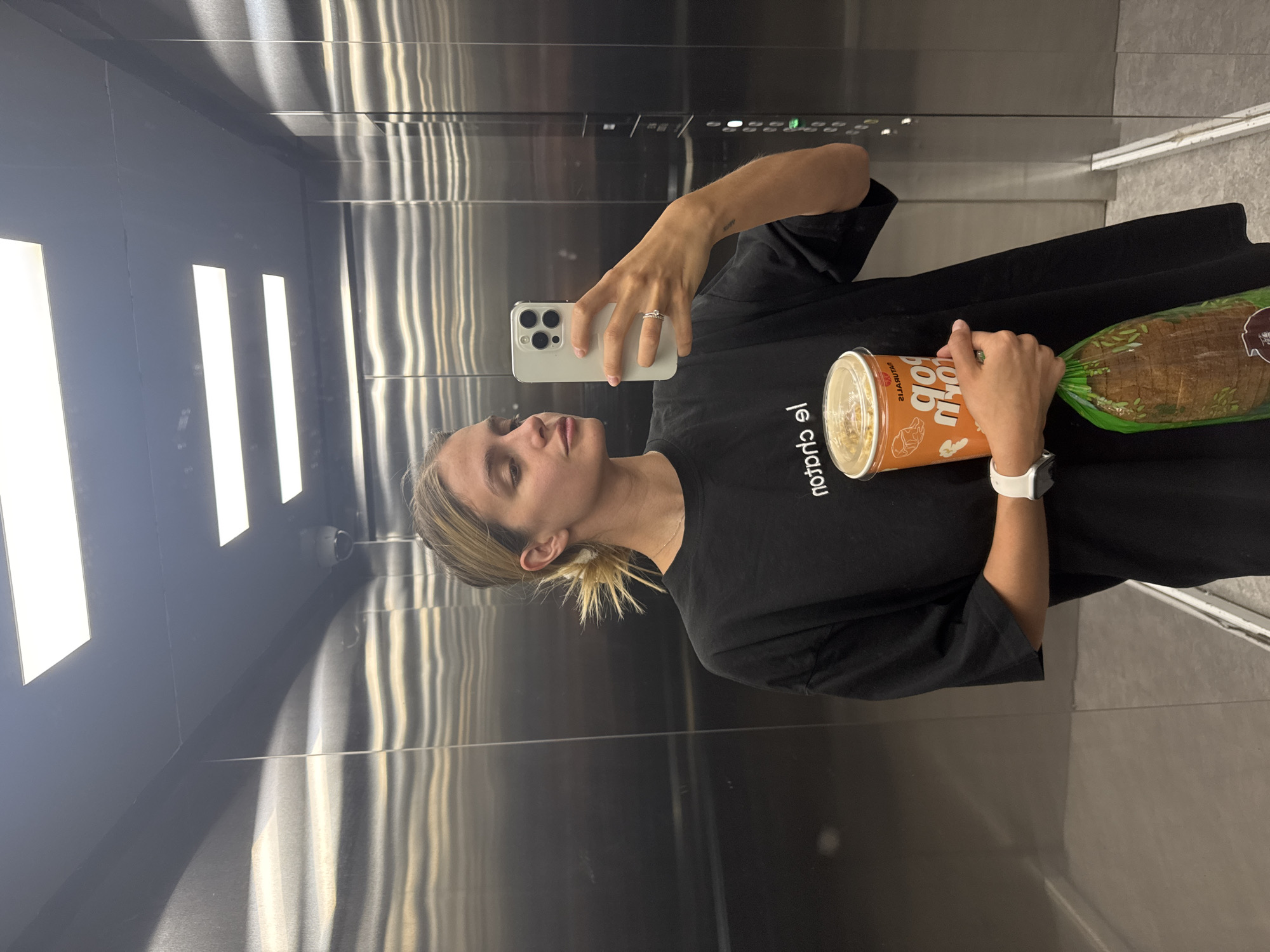 A woman stands in an elevator, holding a container of popcorn and a loaf of bread. She is taking a selfie with a smartphone. The elevator's interior is metallic, with recessed lighting. The location is Unknown, Unknown.