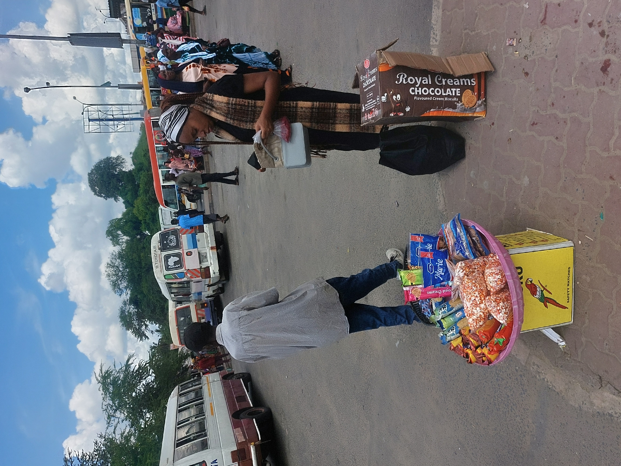 The image depicts a street scene in Dar es-Salaam, Tanzania. There are two vendors: one woman selling clothes and another man selling snacks. The woman is sitting next to a pile of clothes and a box of "Royal Creams Chocolate Flavoured Cream Biscuits". She is holding a small bag and a small, light-colored container. The man is standing with a basket full of various snacks. The basket is being supported by a box for "Safety Matches", and also contains "Marie" biscuits and "Royal Creams" biscuits. In the background, there are several white minibuses (dala-dalas) lined up, and people can be seen walking around. The sky is blue with scattered clouds. The ground appears to be paved with stone.