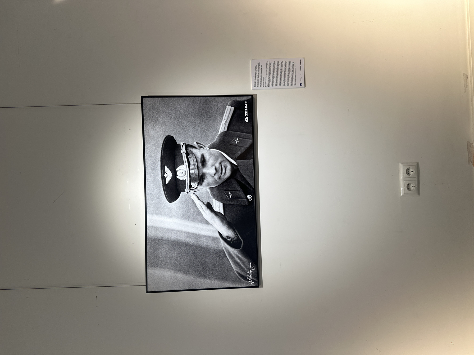 The photograph captures a black and white portrait of Yuri Gagarin, the first human to journey into outer space, in uniform and saluting. The image is framed and hung on a white wall, likely in a museum or gallery, indicated by the informational placard to the right of the photograph. Below the framed photograph, a pair of electrical outlets are visible on the wall. The lighting appears to be artificial, creating a slightly warm glow on the wall, particularly on the right side. The photograph is suspended by thin wires, suggesting a modern exhibition setup. The informational placard contains text in Russian, with logos of organizations such as TASS, ZENIT, and others, further supporting the context of an exhibition in Moscow, Russia. The overall scene is clean and organized, typical of a public exhibition space.