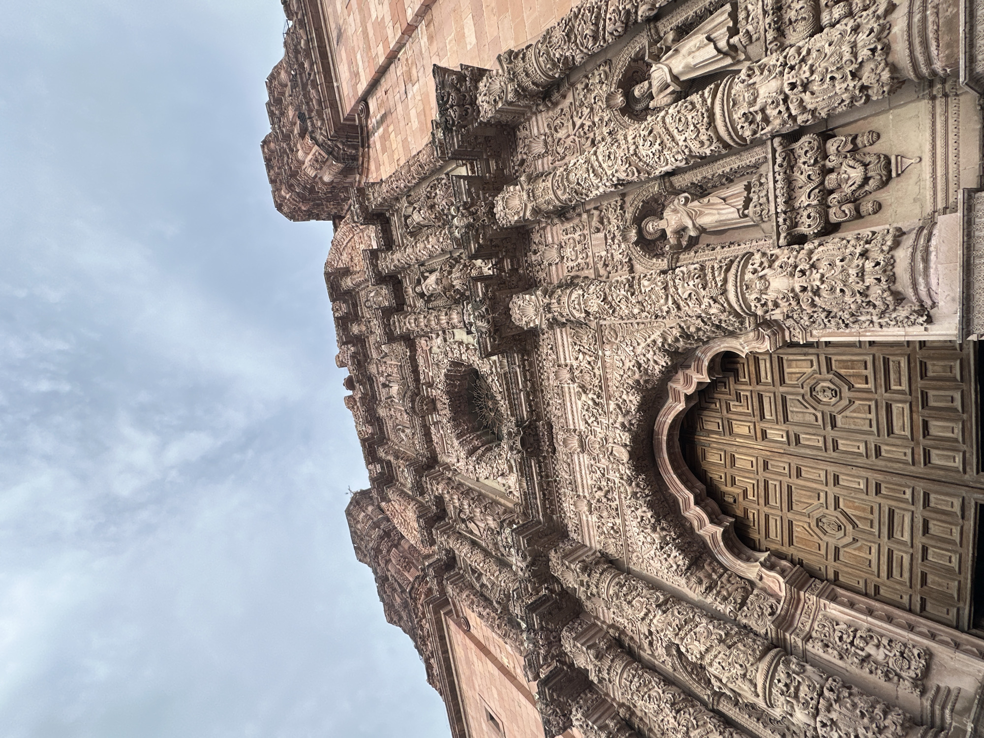 The image shows a low-angle, wide shot of the intricately carved facade of the Zacatecas Cathedral in Zacatecas City, Mexico. The cathedral, constructed of pinkish-red stone, features a highly ornate Baroque style, with detailed sculptures, columns, and decorative motifs covering the entire surface of the visible entrance. Two statues of figures in robes are visible within niches on either side of the main arched entryway, which is filled with a large, multi-paneled wooden door. Above the door, the stonework continues with a dense array of relief carvings. The sky above is overcast with scattered clouds. The perspective emphasizes the grandeur and overwhelming detail of the architecture.