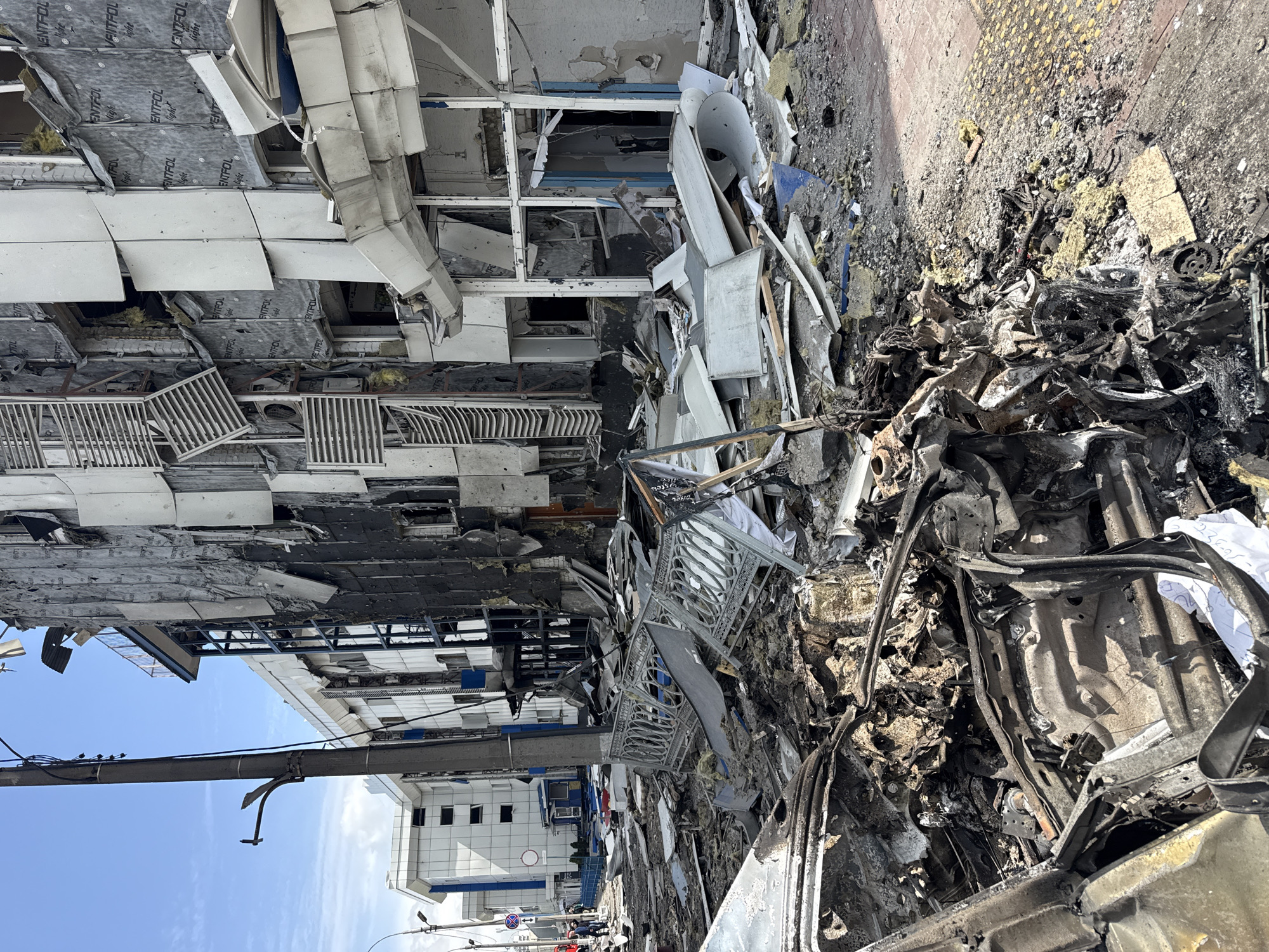The image depicts a scene of devastation, seemingly the aftermath of an explosion or attack. The location appears to be in Kyiv, Ukraine. A multi-story building is heavily damaged, with portions of its facade destroyed or collapsed, revealing interior structures and debris. The ground is covered in rubble, including charred remains of what may have been a vehicle. There are no visible people in the immediate foreground. The sky is partly cloudy. Overall, the scene suggests significant structural damage and destruction.