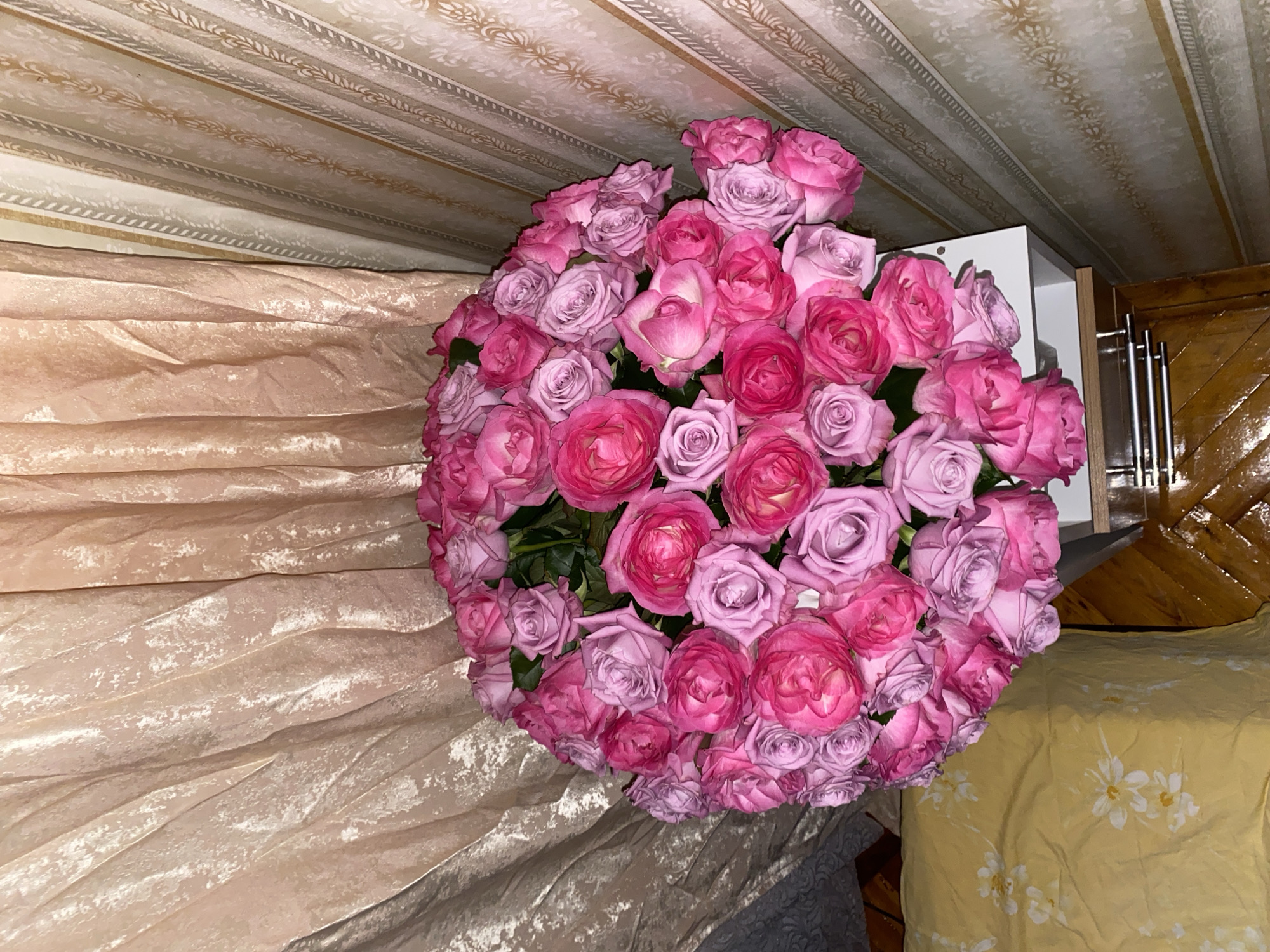 A large, spherical bouquet of pink and lavender roses is positioned on a white surface, likely a table or shelf. The bouquet is densely packed with roses of varying shades, from deep fuchsia to pale lilac. Behind the bouquet, the background features textured curtains with a soft, golden hue and a patterned wallpaper in cream and gold. To the left of the bouquet, a portion of a yellow floral-patterned sheet is visible, suggesting a bedroom setting. The overall impression is one of floral abundance and soft, warm lighting. The image was taken in the city of Lomonosov, Russia.