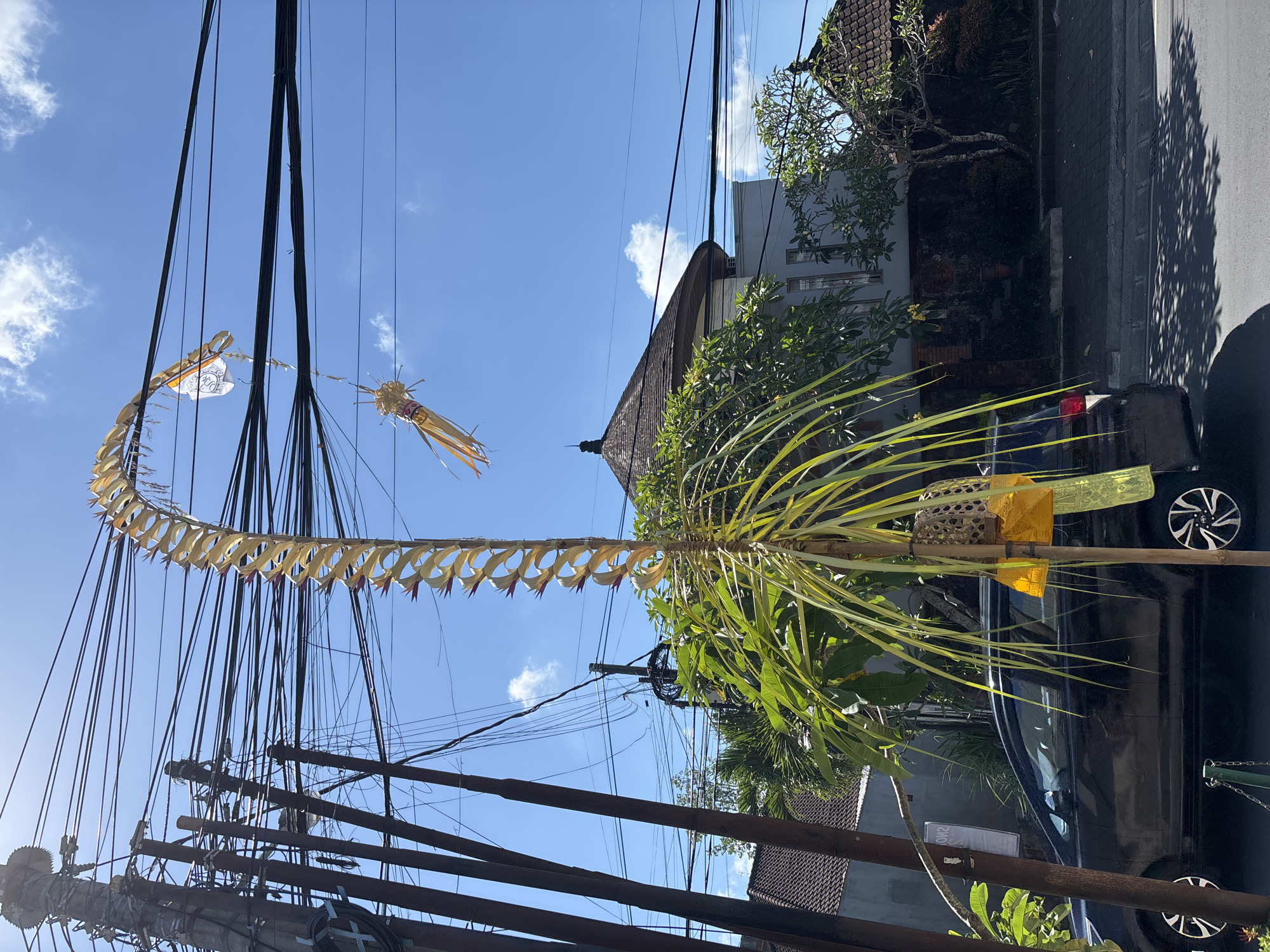 This is a street-level, eye-level shot taken in Kuta Selatan, Indonesia, on a clear, sunny day with scattered white clouds. The foreground is dominated by a tall, decorative bamboo structure adorned with palm leaves and yellow fabric, likely a ceremonial item. To the left, a dark-colored car is parked on the side of a paved road. In the background, houses with traditional tiled roofs and a modern blue-gray building are visible, partially obscured by lush green trees and foliage. A complex network of overhead electrical wires crisscrosses the sky, casting shadows on the ground. Two smaller decorative items, also appearing to be made of woven material and fabric, are suspended from these wires. The overall scene suggests a cultural celebration or event taking place in a tropical urban environment.