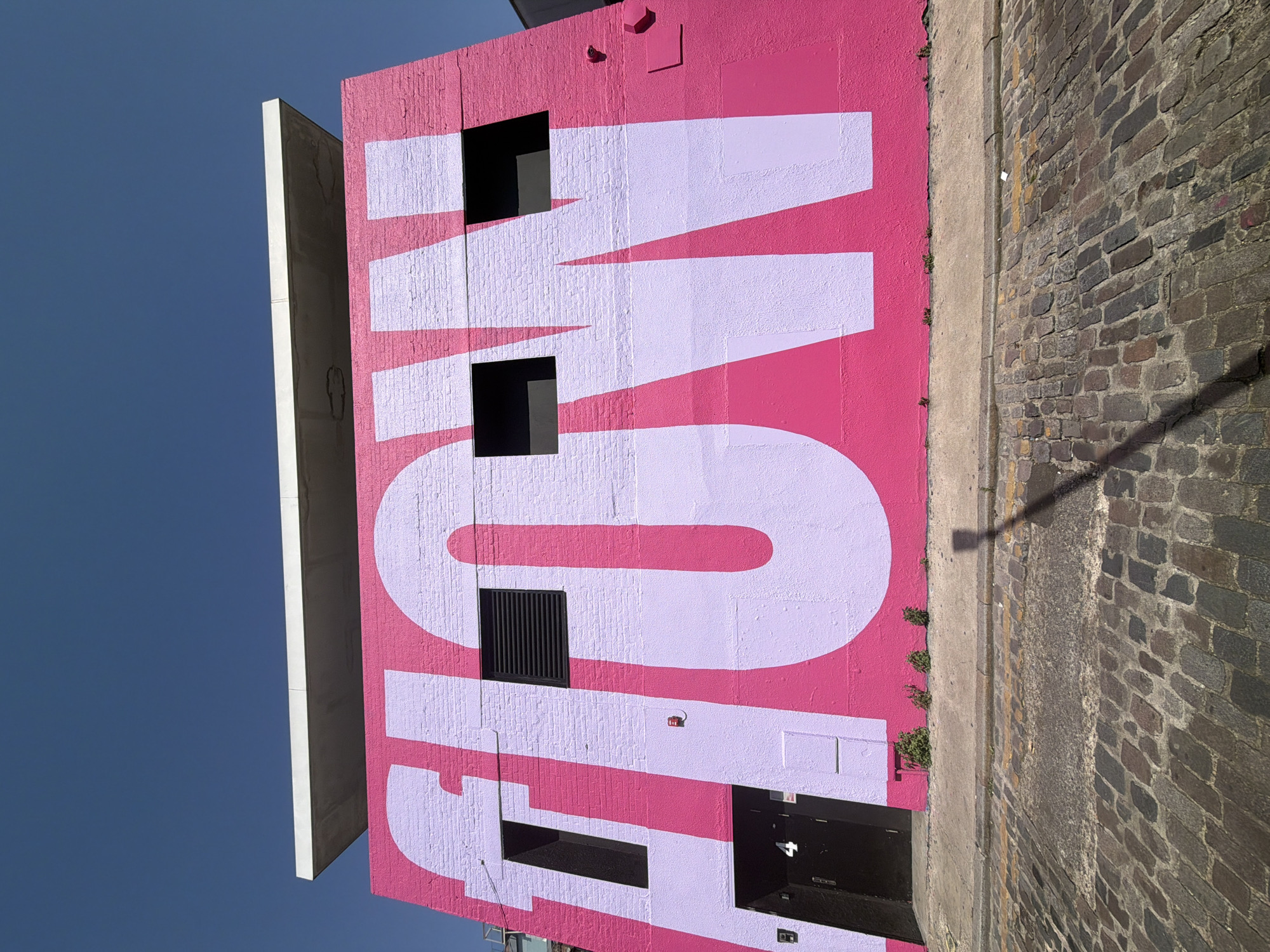 A vertical outdoor shot features the side of a building with a pink facade painted with the large word "FLOW" in a lighter lavender color. The letters are partially interrupted by architectural elements like windows and vents. The sky is visible on the left, and a brick street borders the building on the right. The location is likely in the city of London, United Kingdom.